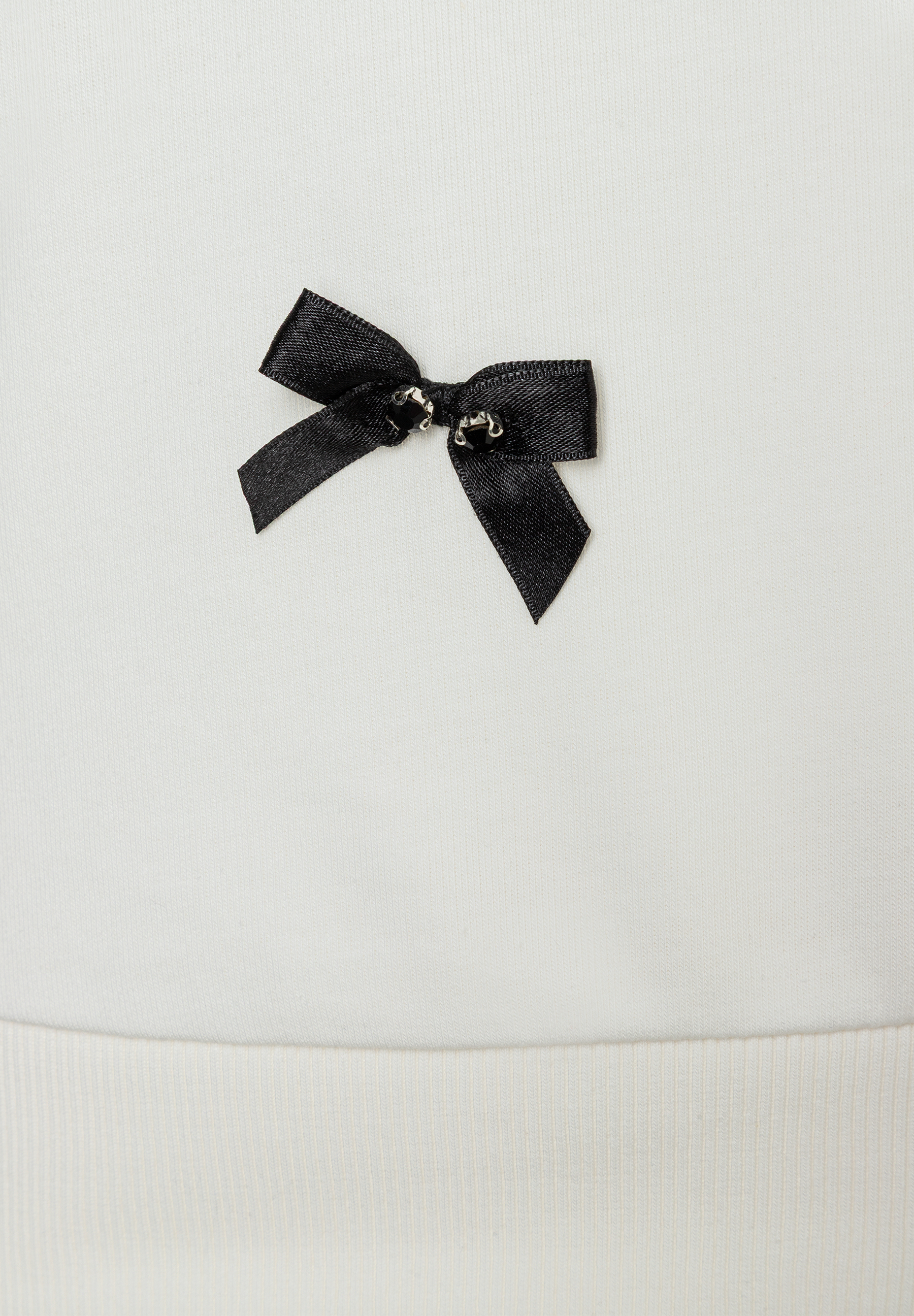 Detailansicht eines weißen MORE & MORE Sweatshirts mit einer kleinen, schwarzen Schleife. Detailansicht eines weißen MORE & MORE Sweatshirts mit einer kleinen, schwarzen Schleife.