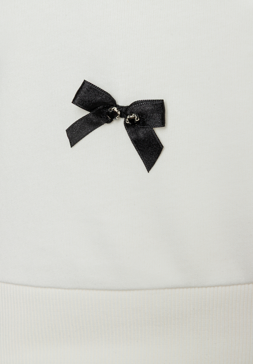 Detailansicht eines weißen MORE & MORE Sweatshirts mit einer kleinen, schwarzen Schleife. Detailansicht eines weißen MORE & MORE Sweatshirts mit einer kleinen, schwarzen Schleife.