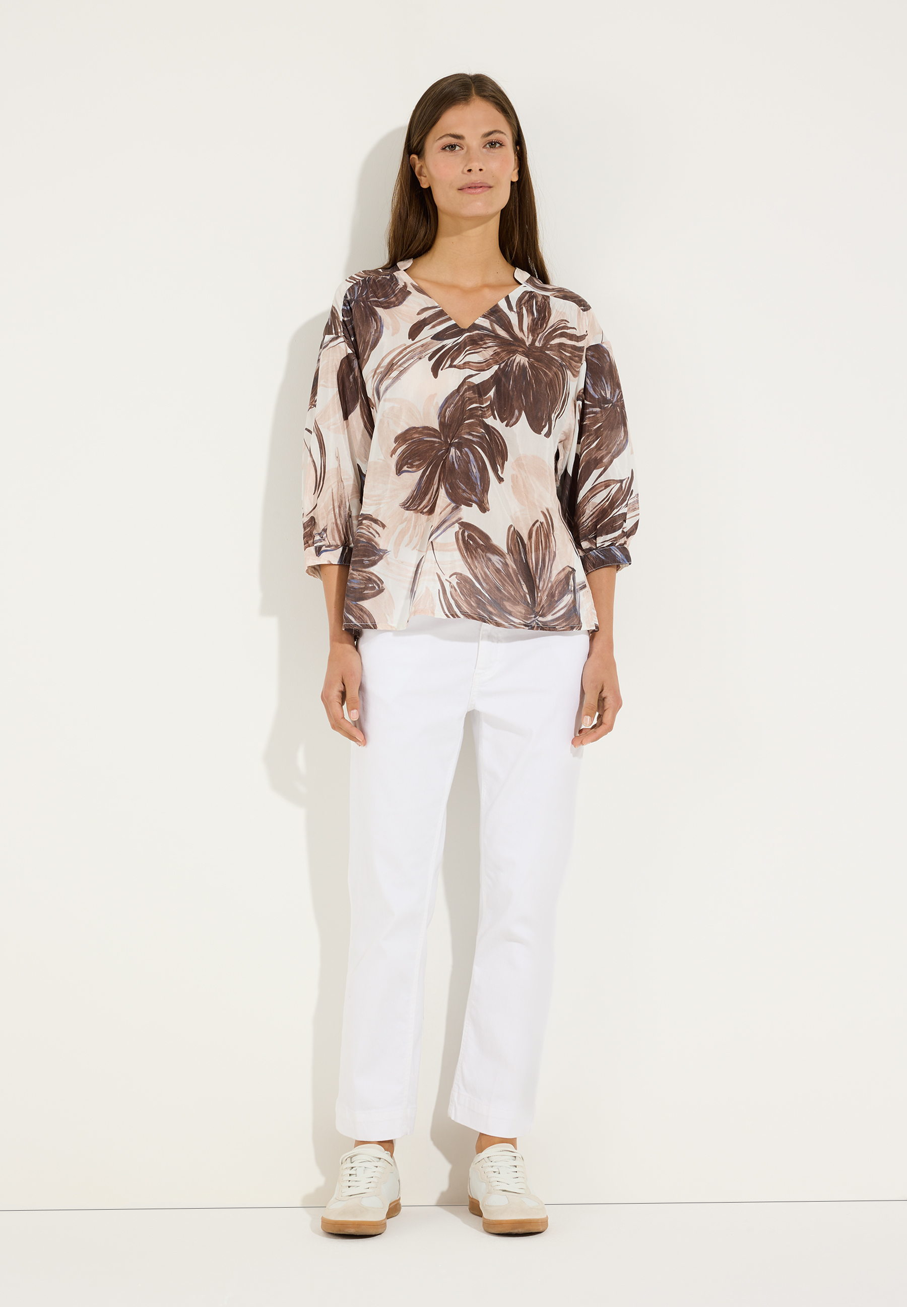 MORE & MORE Damen-Bluse mit V-Ausschnitt, 3/4-Ärmeln und braunem Blumenmuster, kombiniert mit weißer Hose und beigen Sneakern. MORE & MORE Damen-Bluse mit V-Ausschnitt, 3/4-Ärmeln und braunem Blumenmuster, kombiniert mit weißer Hose und beigen Sneakern.
