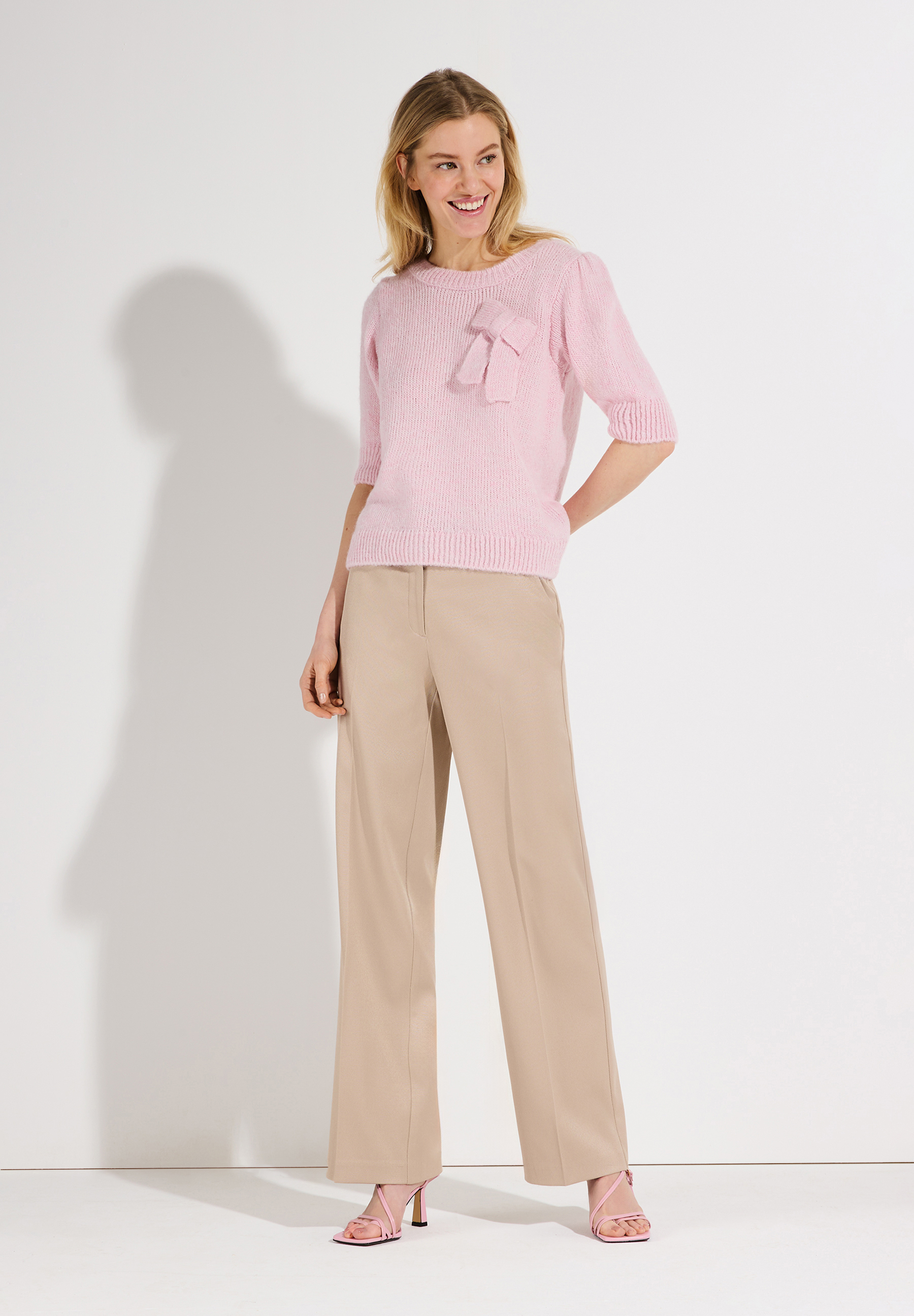 Eine Frau trägt einen MORE & MORE Strickpullover mit Schleife und eine Hose in hellem Beige.