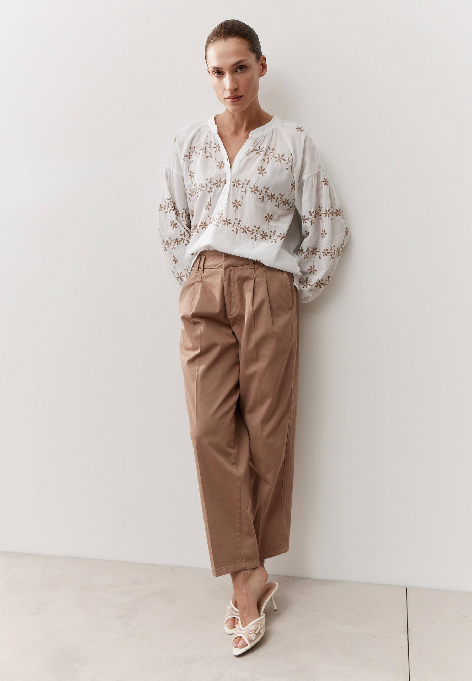 Eine Frau in MORE & MORE Oberteilen, mit brauner Hose und weißen Schuhen vor einer weißen Wand.