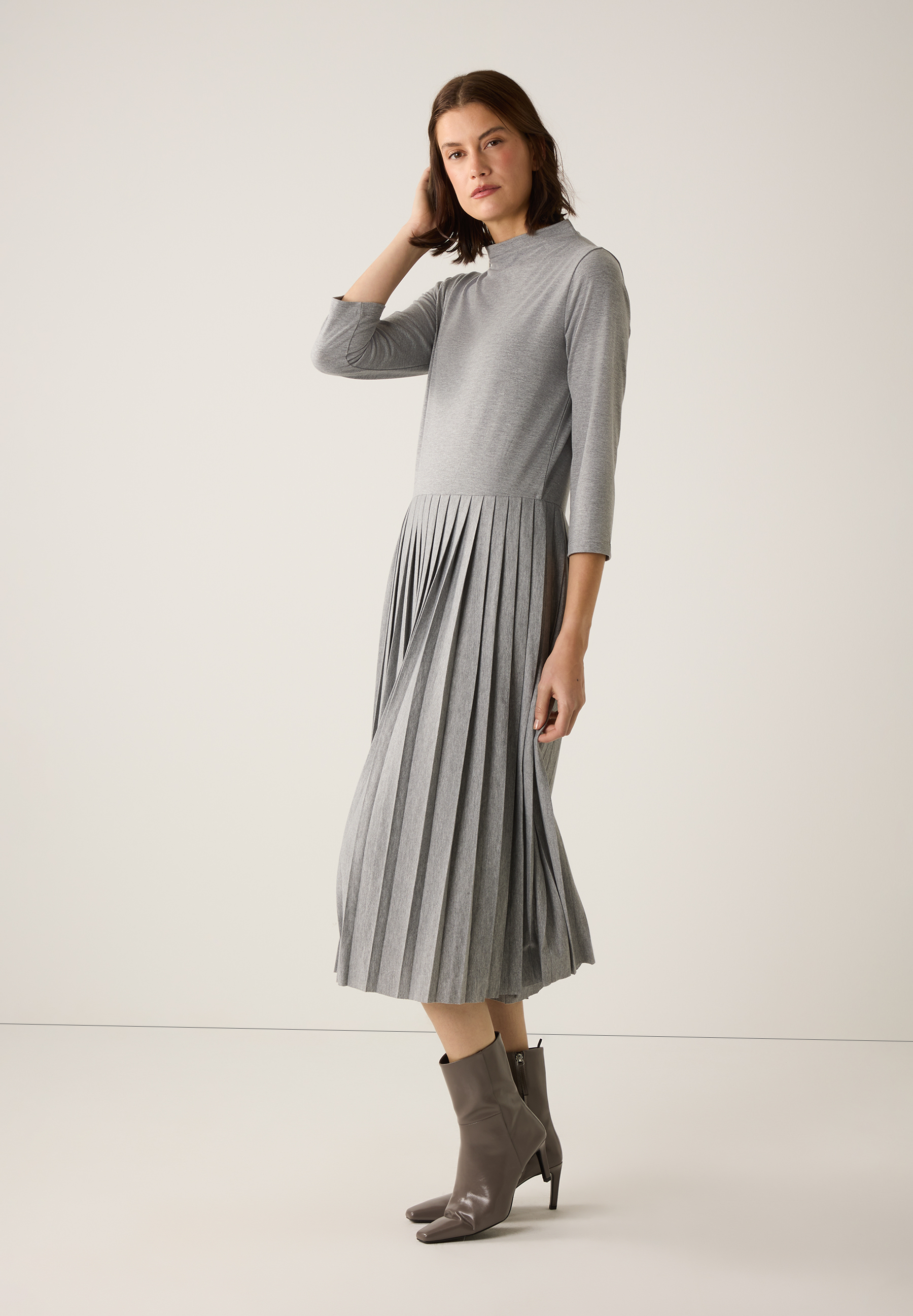 Eine Frau mit grauem MORE & MORE Kleid mit Jersey-Oberteil und Faltenrock, dazu passende Stiefeletten. Eine Frau mit grauem MORE & MORE Kleid mit Jersey-Oberteil und Faltenrock, dazu passende Stiefeletten.