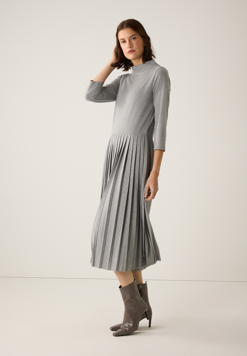 Eine Frau mit grauem MORE & MORE Kleid mit Jersey-Oberteil und Faltenrock, dazu passende Stiefeletten. Eine Frau mit grauem MORE & MORE Kleid mit Jersey-Oberteil und Faltenrock, dazu passende Stiefeletten.