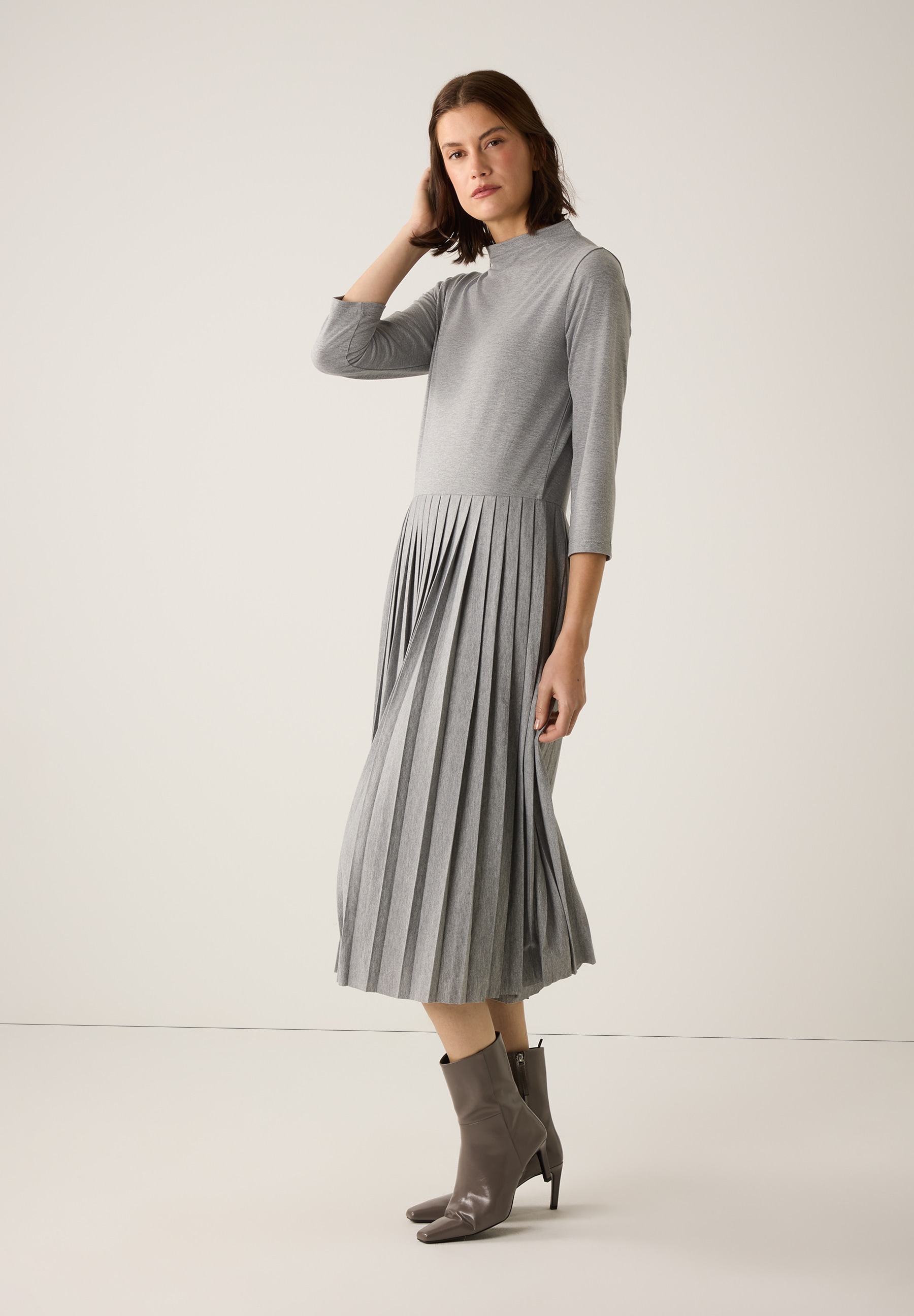 Eine Frau mit grauem MORE & MORE Kleid mit Jersey-Oberteil und Faltenrock, dazu passende Stiefeletten. Eine Frau mit grauem MORE & MORE Kleid mit Jersey-Oberteil und Faltenrock, dazu passende Stiefeletten.