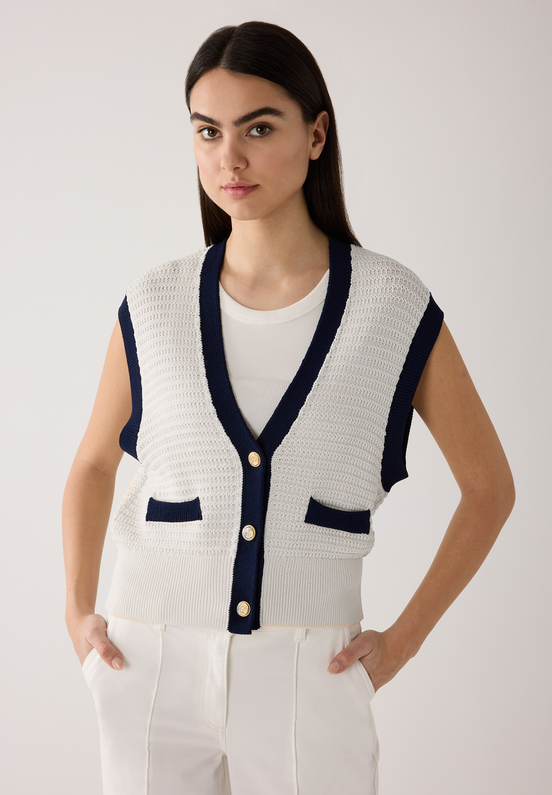 Eine Frau präsentiert einen weißen Strukturstrick-Cardigan von MORE & MORE mit marineblauen Akzenten und goldenen Knöpfen.