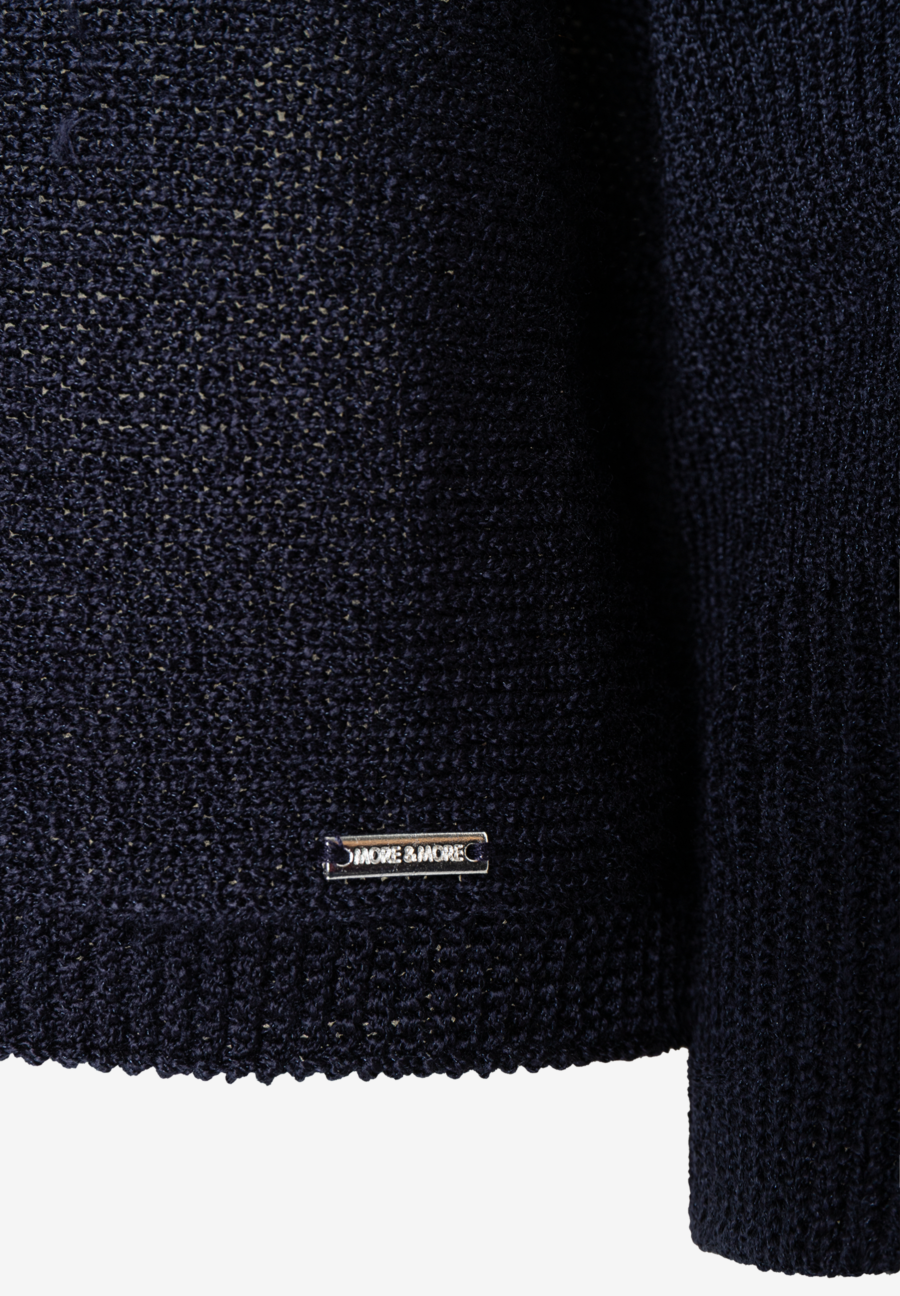 Nahaufnahme eines dunkelblauen Strickoberteils von MORE & MORE mit glänzender Optik und Metallemblem der Marke. Nahaufnahme eines dunkelblauen Strickoberteils von MORE & MORE mit glänzender Optik und Metallemblem der Marke.
