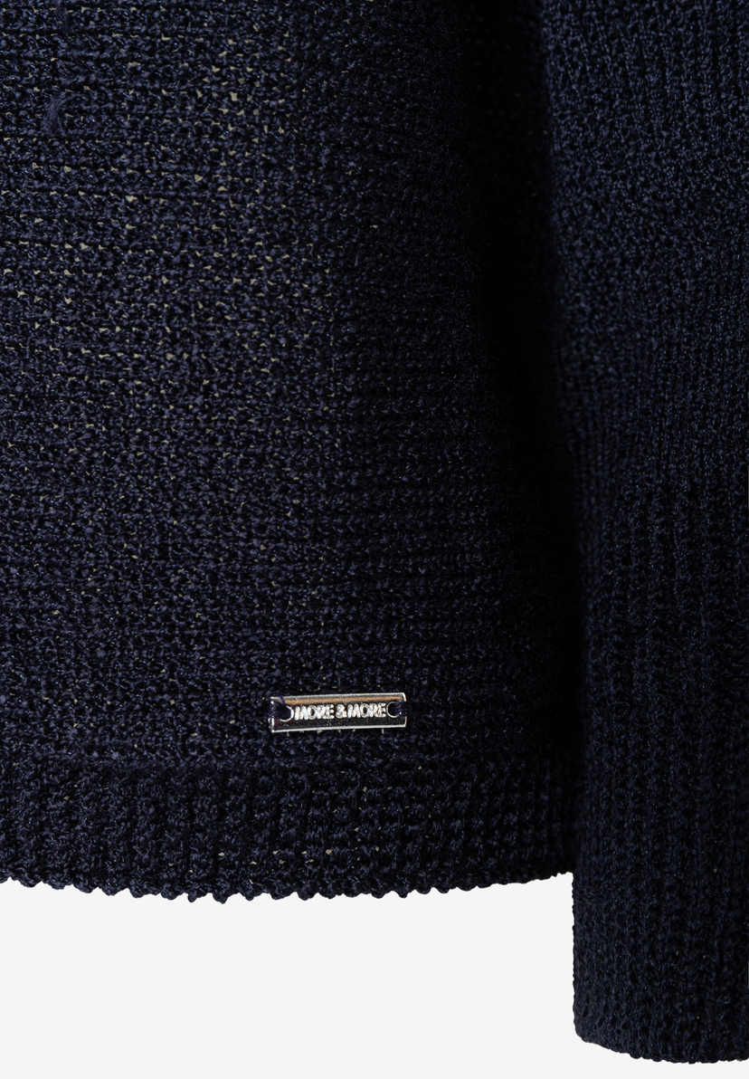 Nahaufnahme eines dunkelblauen Strickoberteils von MORE & MORE mit glänzender Optik und Metallemblem der Marke. Nahaufnahme eines dunkelblauen Strickoberteils von MORE & MORE mit glänzender Optik und Metallemblem der Marke.