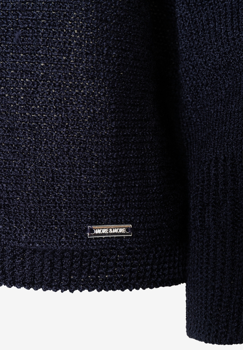 Nahaufnahme eines dunkelblauen Strickoberteils von MORE & MORE mit glänzender Optik und Metallemblem der Marke. Nahaufnahme eines dunkelblauen Strickoberteils von MORE & MORE mit glänzender Optik und Metallemblem der Marke.