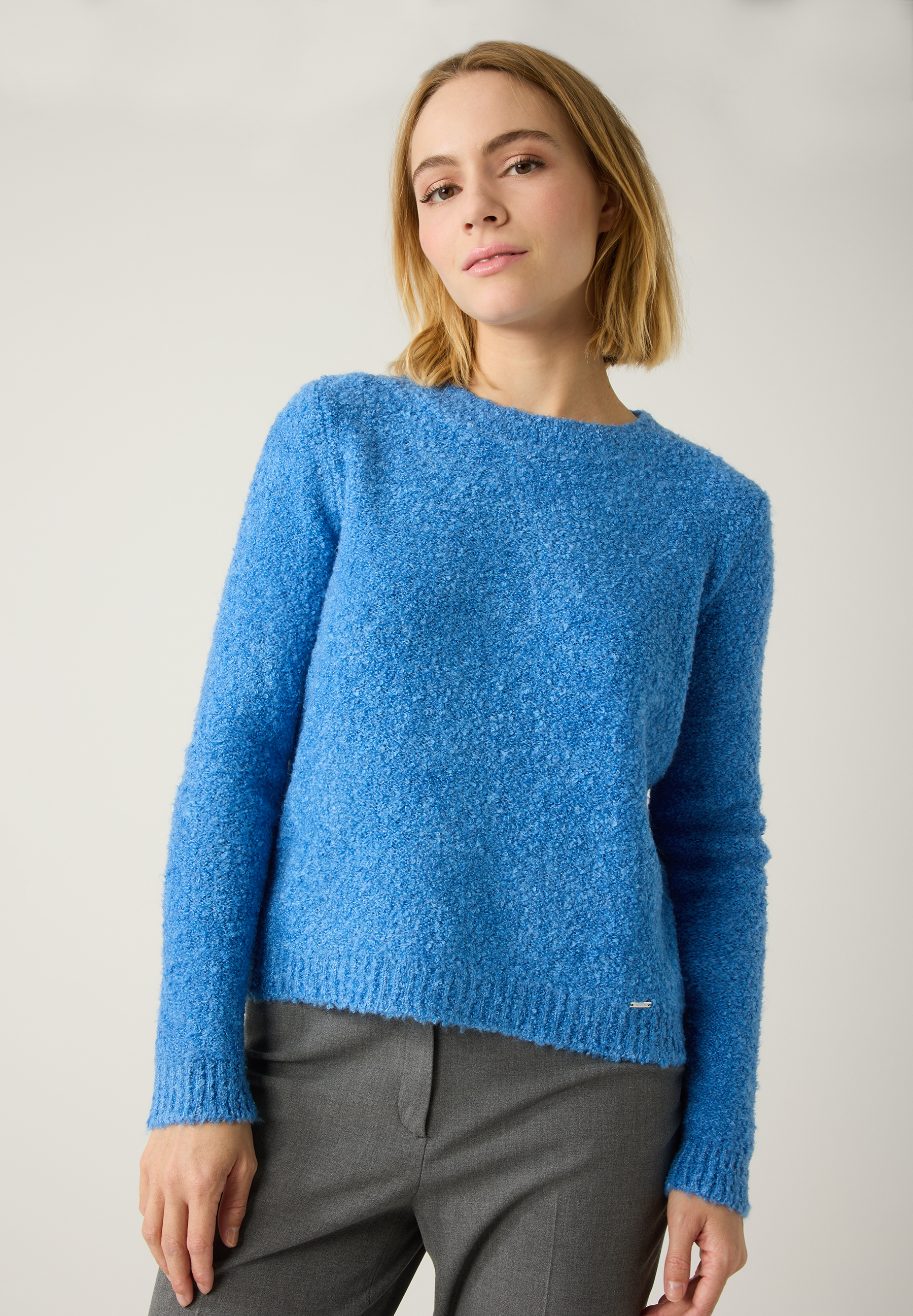 Eine Frau trägt einen blauen Strickpullover von MORE & MORE, kombiniert mit einer grauen Hose vor einem neutralen Hintergrund.