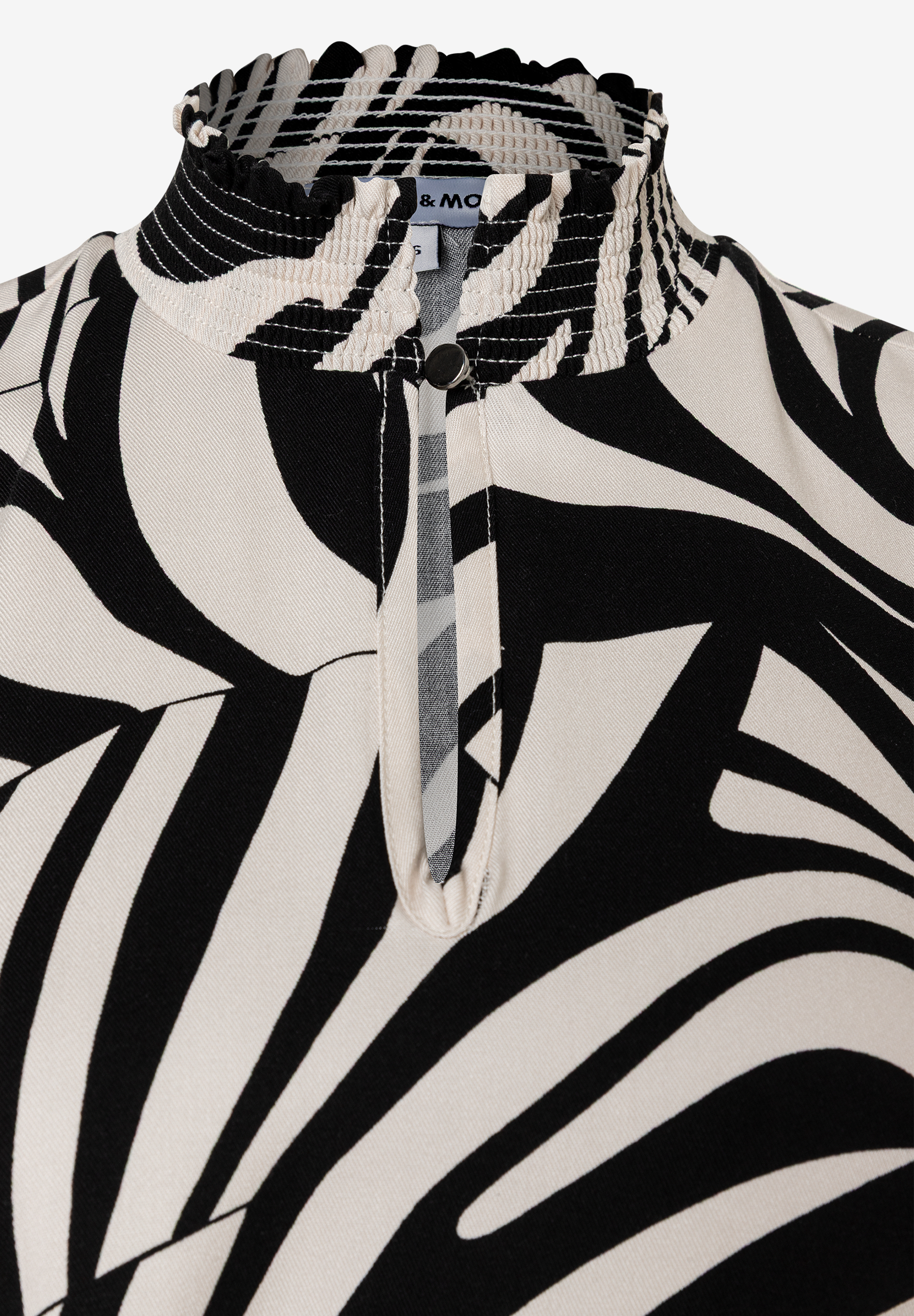 Nahaufnahme eines MORE & MORE Oberteils mit Zebra-Muster, Kräuselkragen und Knopfdetail. Nahaufnahme eines MORE & MORE Oberteils mit Zebra-Muster, Kräuselkragen und Knopfdetail.