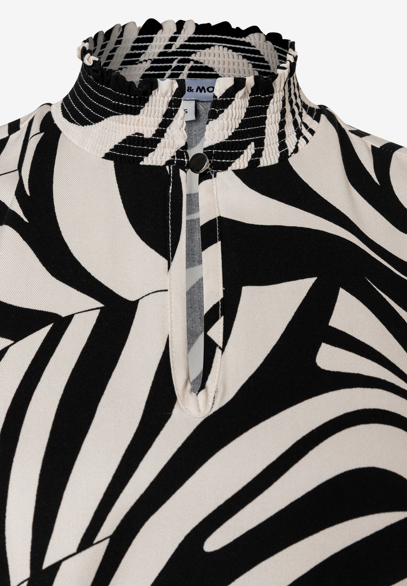 Nahaufnahme eines MORE & MORE Oberteils mit Zebra-Muster, Kräuselkragen und Knopfdetail. Nahaufnahme eines MORE & MORE Oberteils mit Zebra-Muster, Kräuselkragen und Knopfdetail.