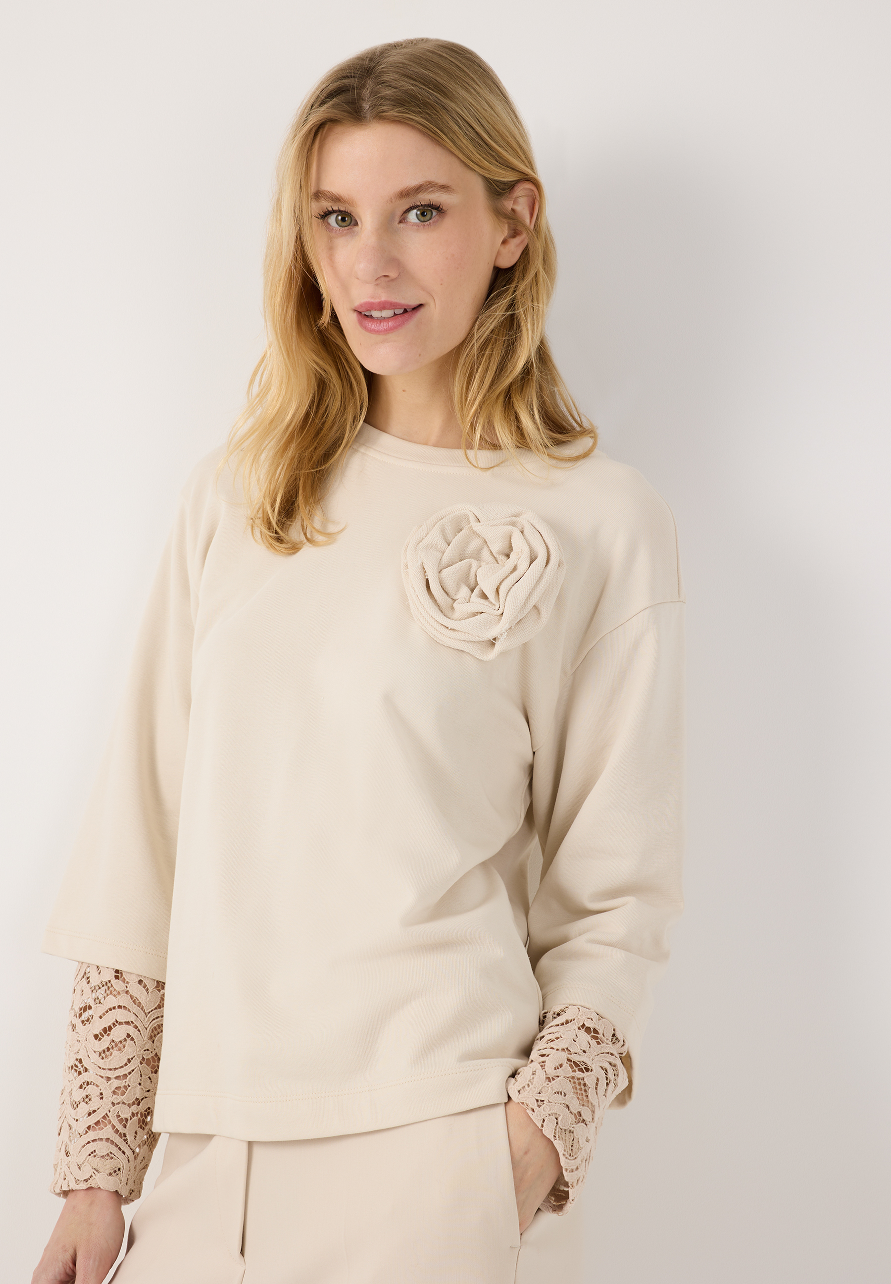 MORE & MORE Sweatshirt in Beige, verziert mit einer cremefarbenen Blütenapplikation auf der Brust und Spitzenmanschetten.