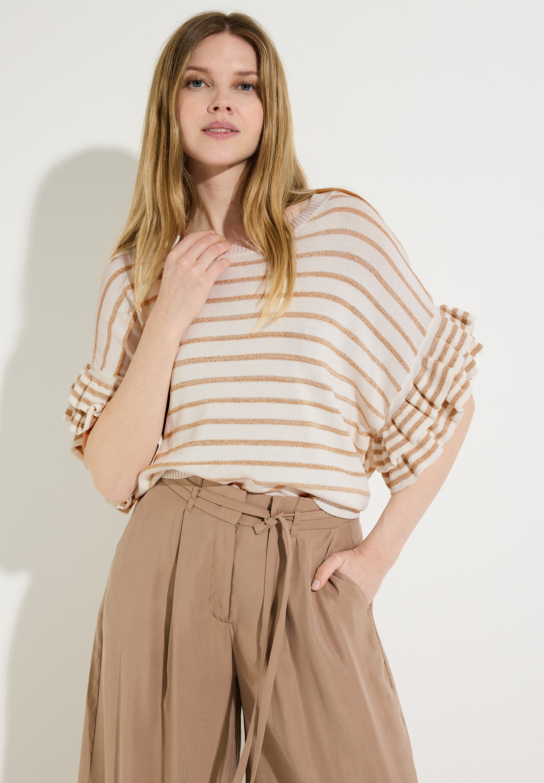 Eine Frau trägt ein MORE & MORE Strickoberteil mit beigen Streifen und Rüschenärmeln, kombiniert mit einer lockeren, braunen Hose.
