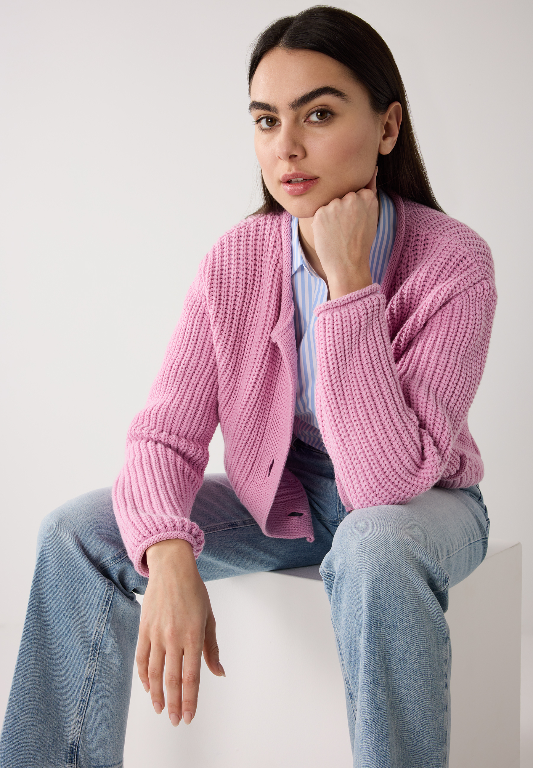 Eine Frau sitzt mit einem MORE & MORE Oberteil aus Rippstrick und Jeans vor einem weißen Hintergrund. Eine Frau sitzt mit einem MORE & MORE Oberteil aus Rippstrick und Jeans vor einem weißen Hintergrund.