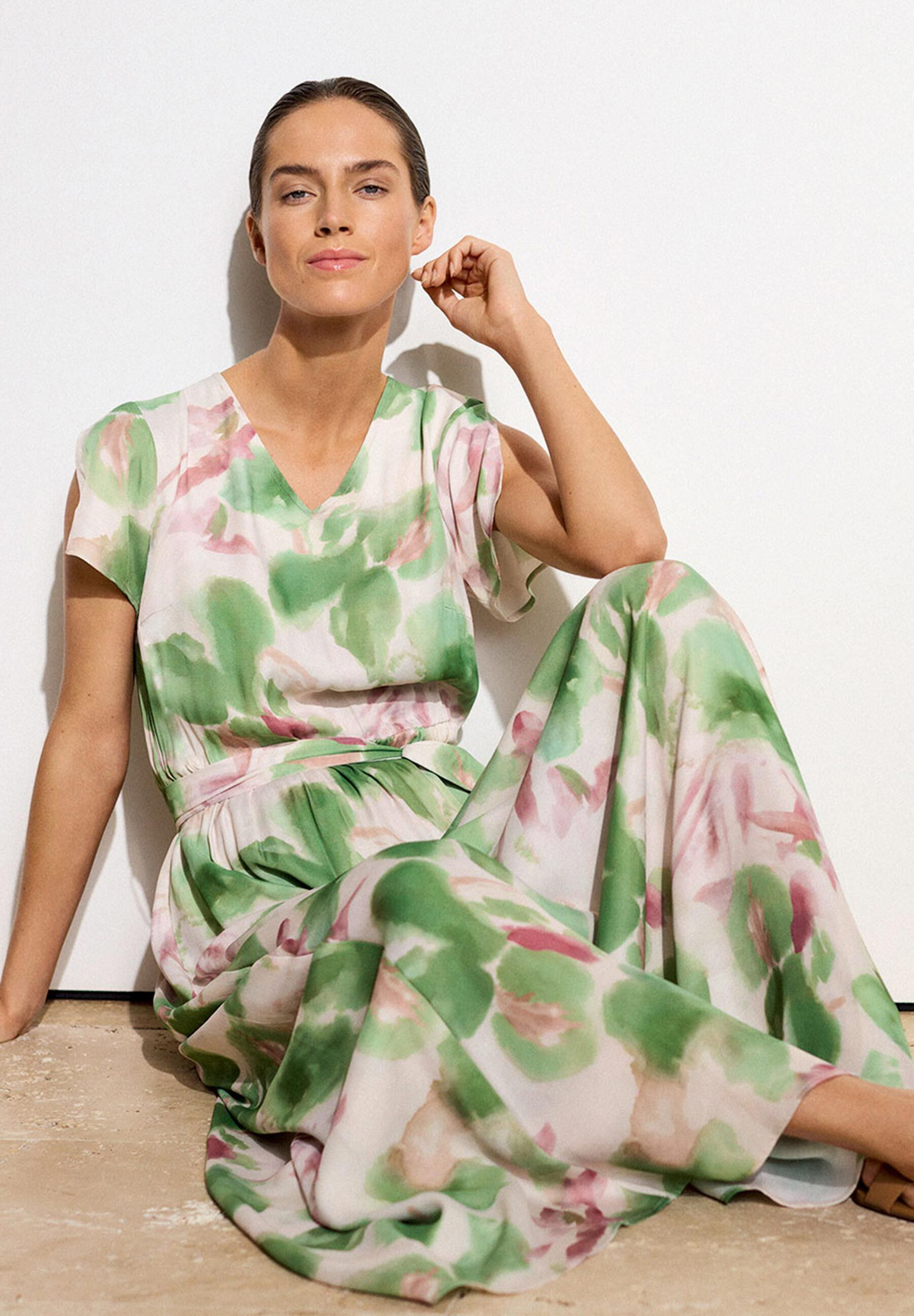 Eine Frau in einem MORE & MORE-Oberteil mit grün-rosa Blätterdesign vor einer weißen Wand.