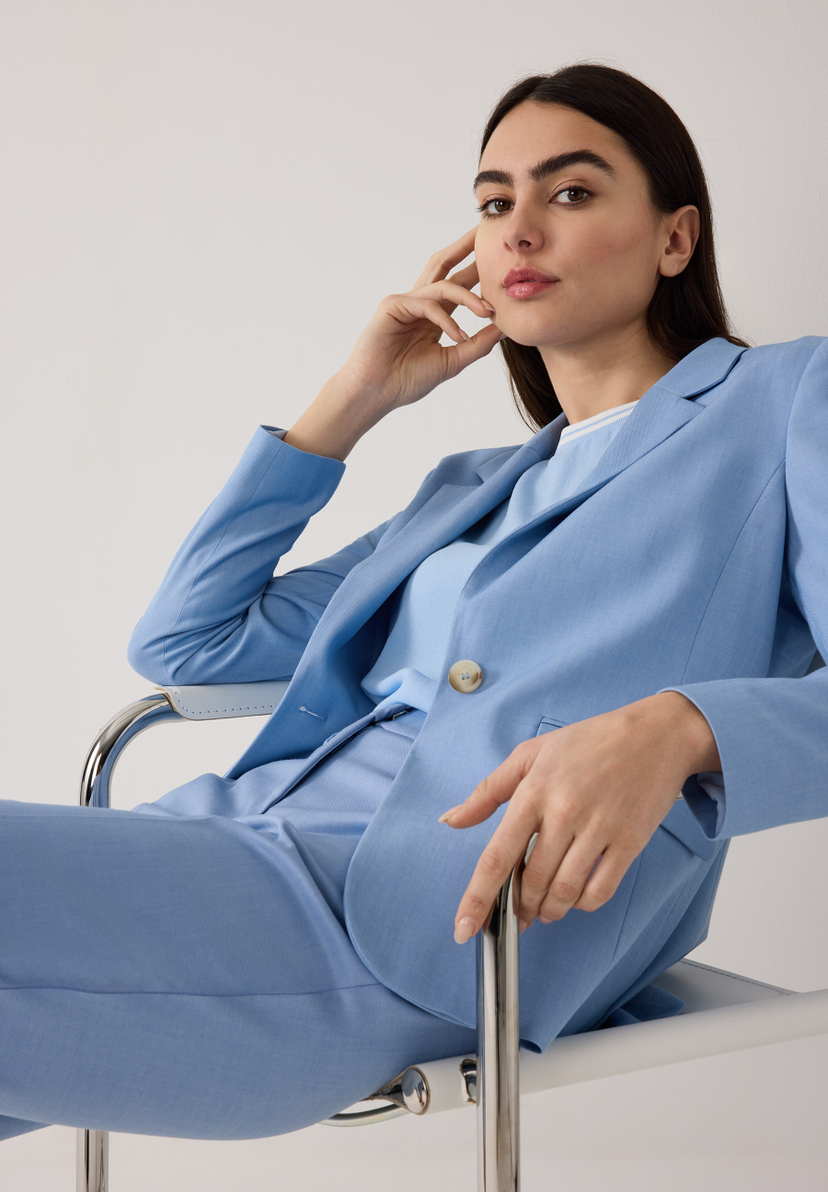Eine Frau in einem blauen MORE & MORE Blazer mit Webware und Stretch sitzt auf einem Stuhl. Eine Frau in einem blauen MORE & MORE Blazer mit Webware und Stretch sitzt auf einem Stuhl.