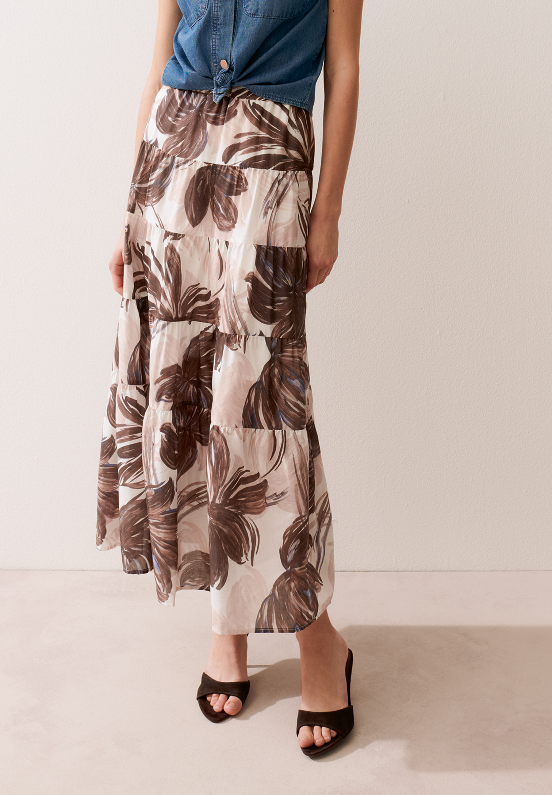 Eine Frau trägt einen MORE & MORE Rock mit Blumenmuster, ein Jeanshemd und schwarze Sandalen.