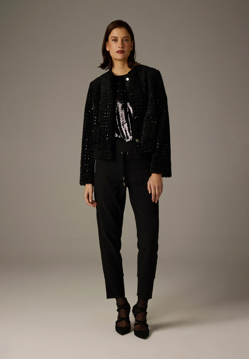 Eine Frau trägt einen schwarzen MORE & MORE Blazer, ein glitzerndes Oberteil und eine Hose vor einem grauen Hintergrund. Eine Frau trägt einen schwarzen MORE & MORE Blazer, ein glitzerndes Oberteil und eine Hose vor einem grauen Hintergrund.