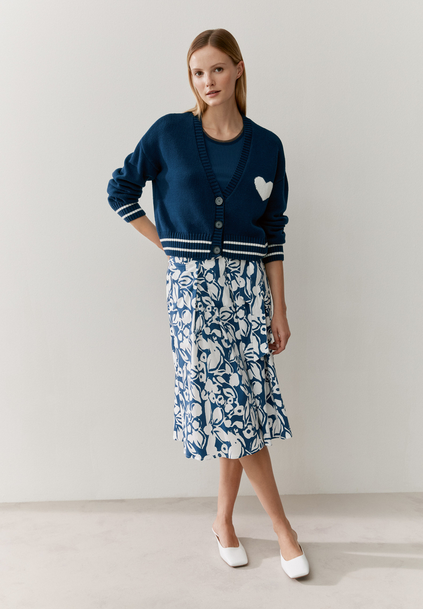 Eine Frau in einem MORE & MORE Strick-Cardigan mit Herz und einem blau-weißen Rock. Eine Frau in einem MORE & MORE Strick-Cardigan mit Herz und einem blau-weißen Rock.