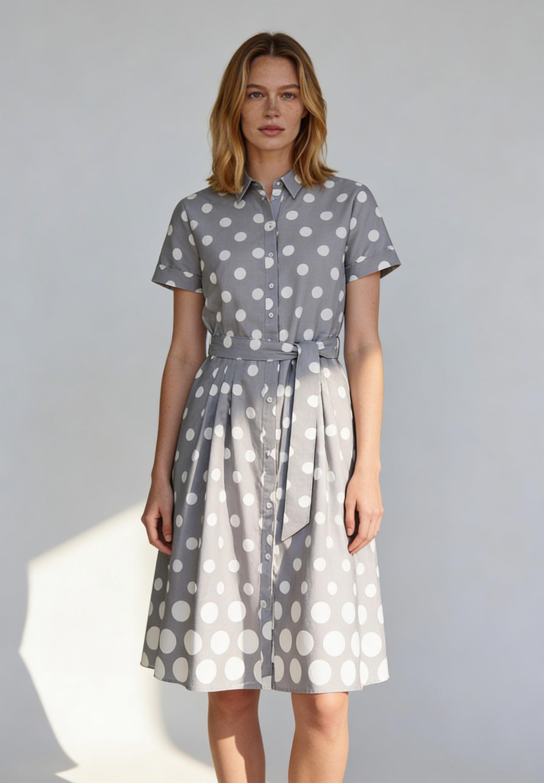 Ein Model trägt ein graues, gepunktetes Kleid von MORE & MORE mit einem Stoffgürtel. Ein Model trägt ein graues, gepunktetes Kleid von MORE & MORE mit einem Stoffgürtel.