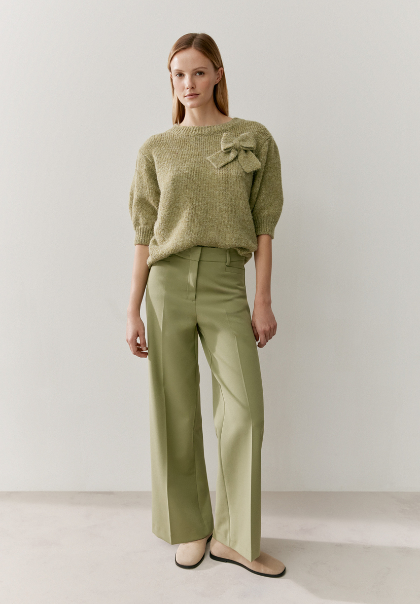 Eine Frau trägt einen grünen MORE & MORE Strickpullover mit Schleife und eine passende Hose. Eine Frau trägt einen grünen MORE & MORE Strickpullover mit Schleife und eine passende Hose.