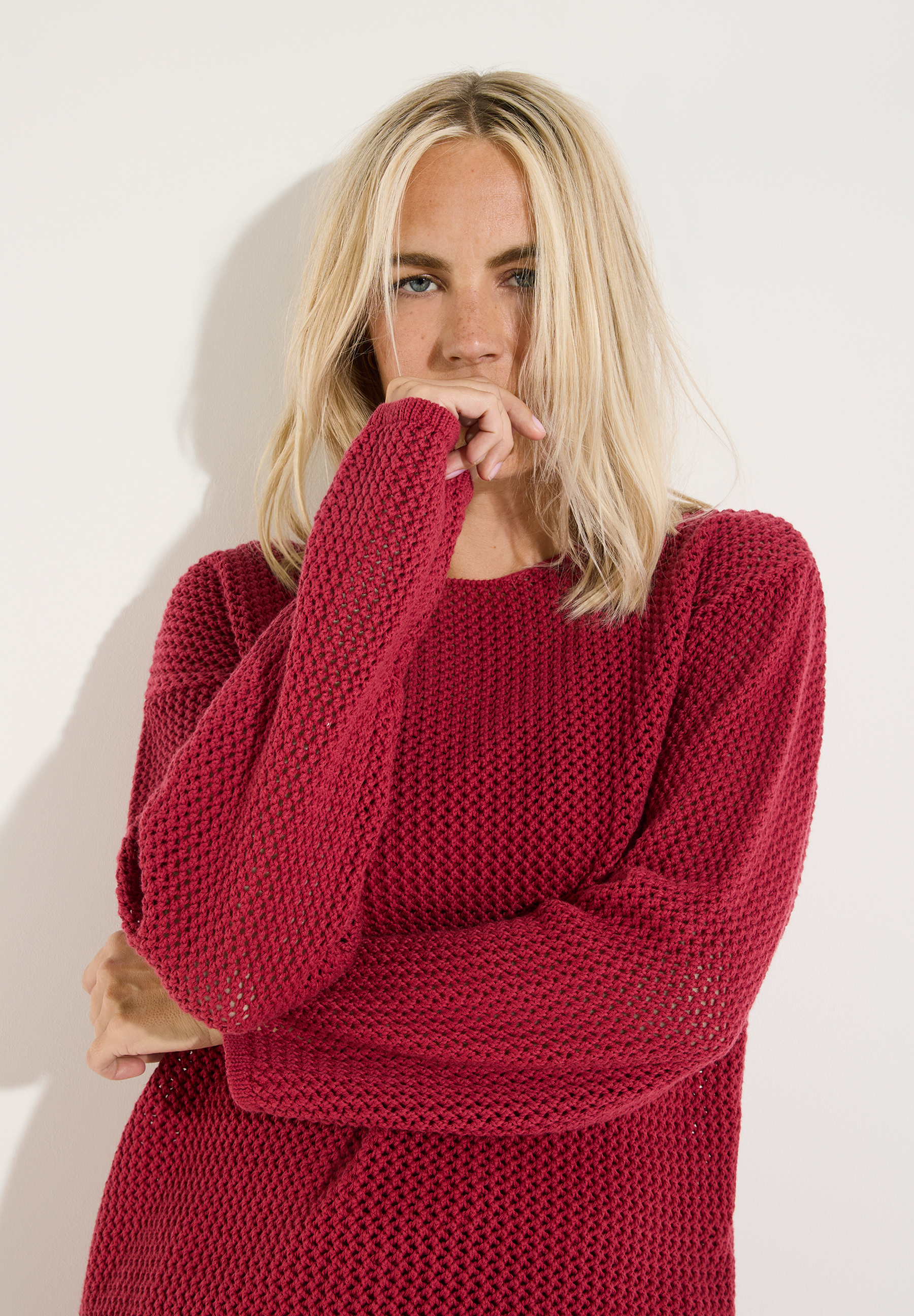 MORE & MORE Pullover in Rot, getragen von einer blonden Frau, die ihre Hand bedeckt und in die Kamera blickt.
