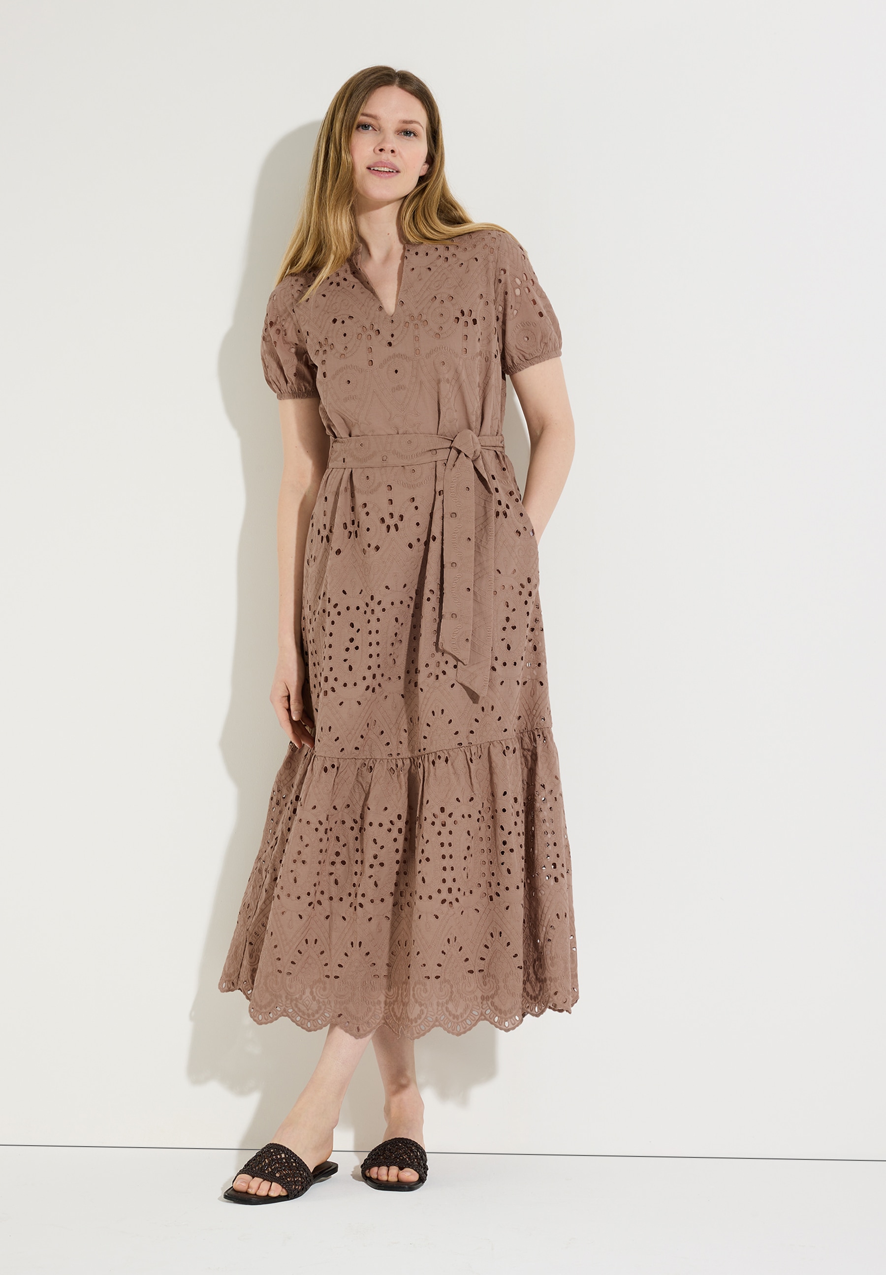 Eine Frau trägt ein MORE & MORE-Kleid aus Spitze mit Gürtel. Das Oberteil ist leicht transparent und hautfreundlich.
