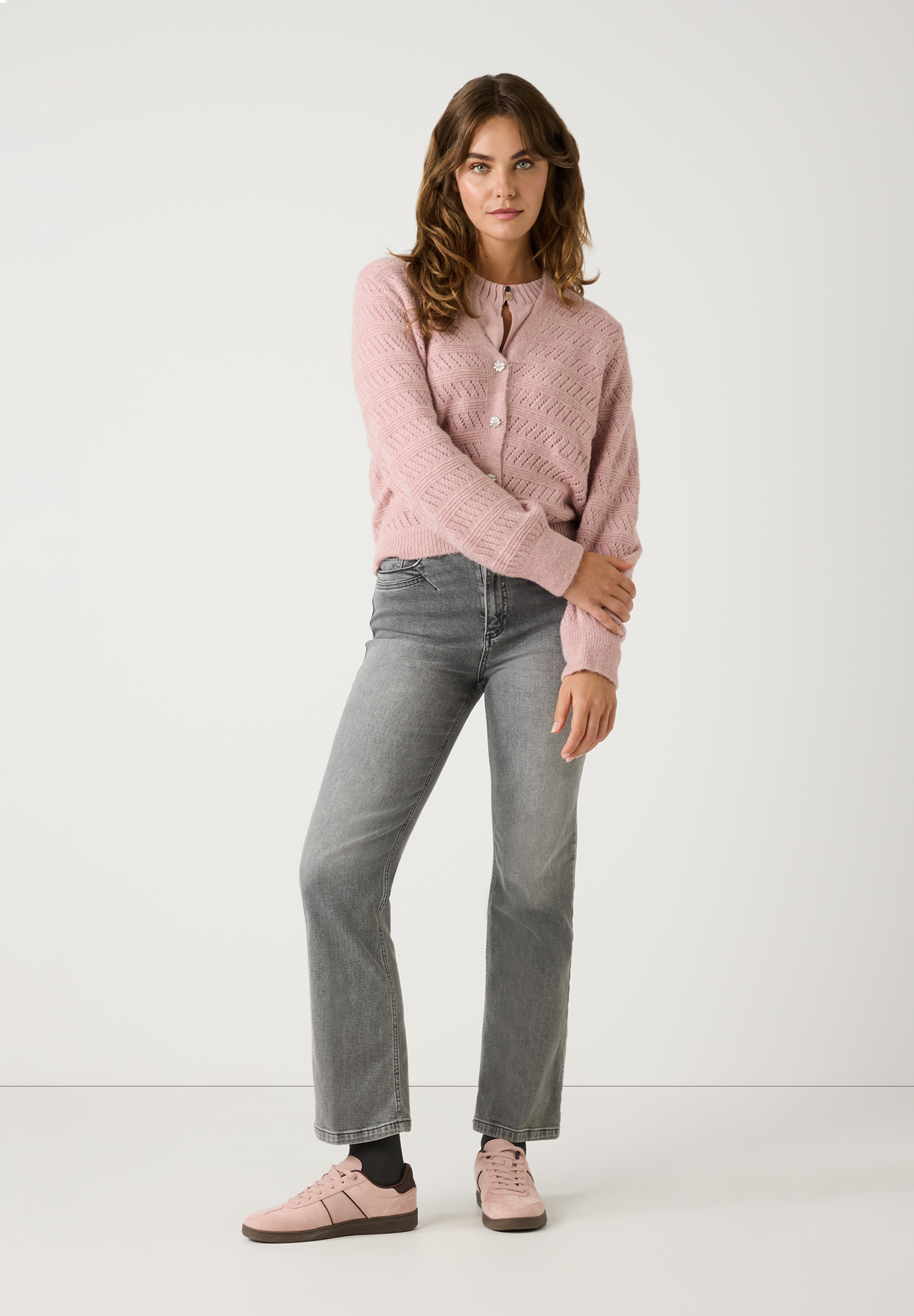 Eine Frau trägt einen MORE & MORE Lochstrick-Cardigan in Altrosa, graue Jeans und rosa Sneakers vor einem weißen Hintergrund. Eine Frau trägt einen MORE & MORE Lochstrick-Cardigan in Altrosa, graue Jeans und rosa Sneakers vor einem weißen Hintergrund.