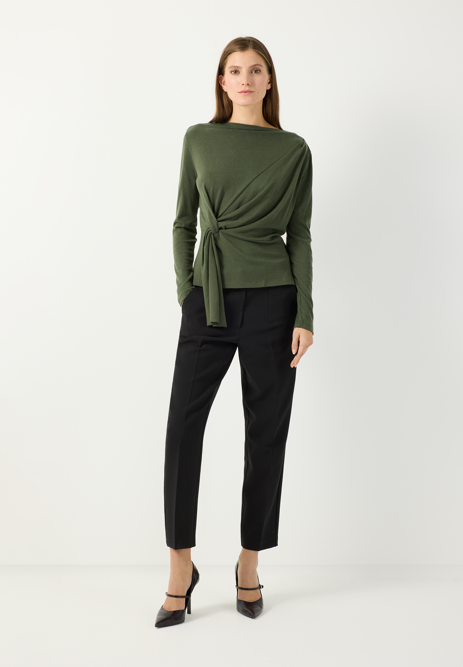 MORE & MORE Jersey-Oberteil in Moosgrün mit Wickeldetail und Bindeknoten. Model trägt das weiche Langarm-Shirt mit U-Boot-Ausschnitt zur schwarzen Hose.