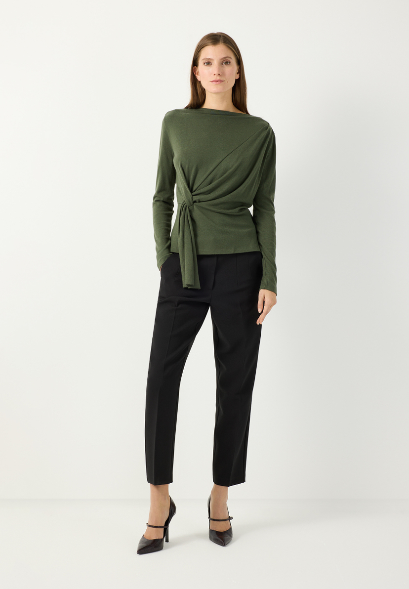 MORE & MORE Jersey-Oberteil in Moosgrün mit Wickeldetail und Bindeknoten. Model trägt das weiche Langarm-Shirt mit U-Boot-Ausschnitt zur schwarzen Hose.