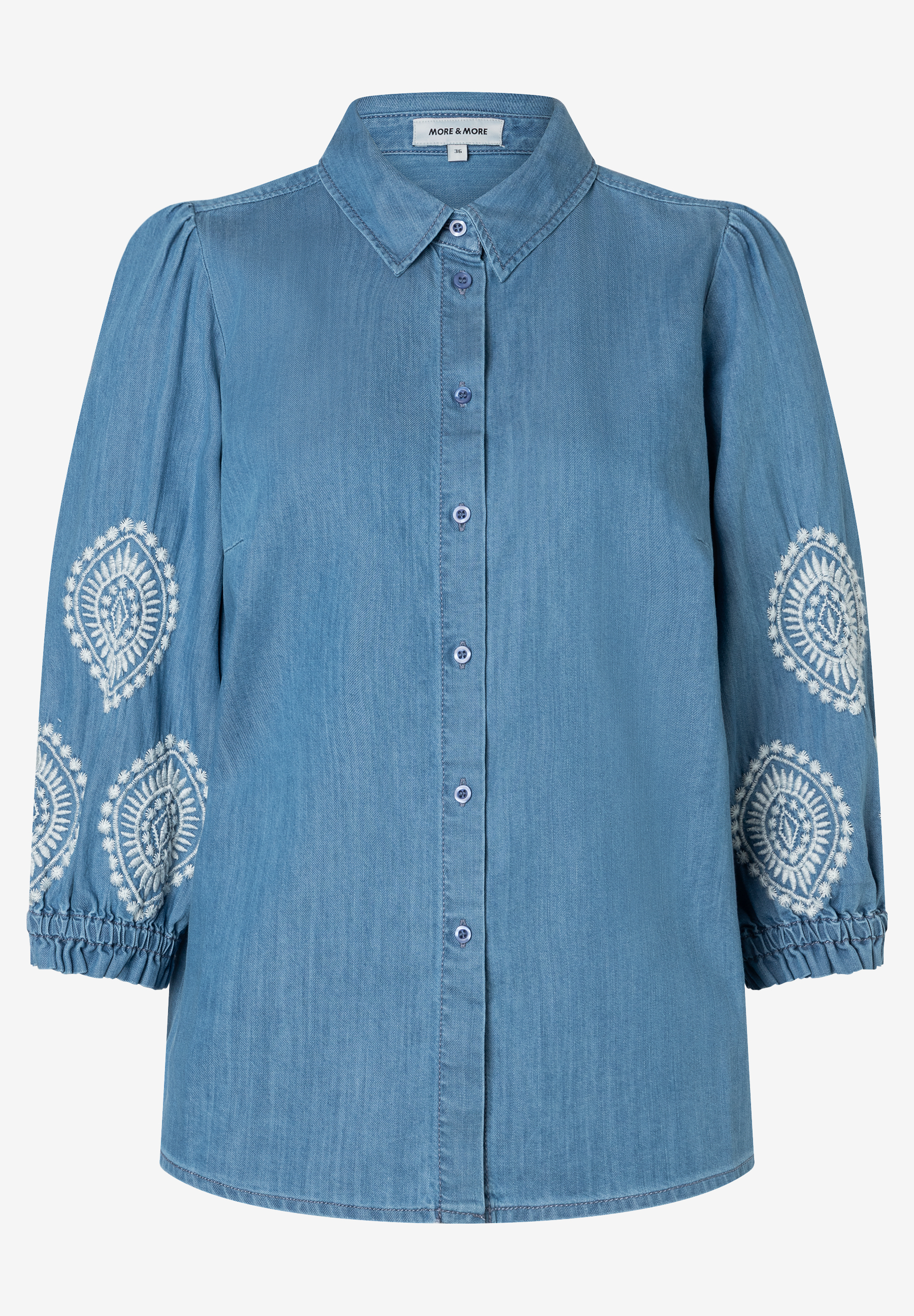 Eine hellblaue Denim-Bluse von MORE & MORE mit Knopfleiste und weißen Stickereien auf den Ärmeln.