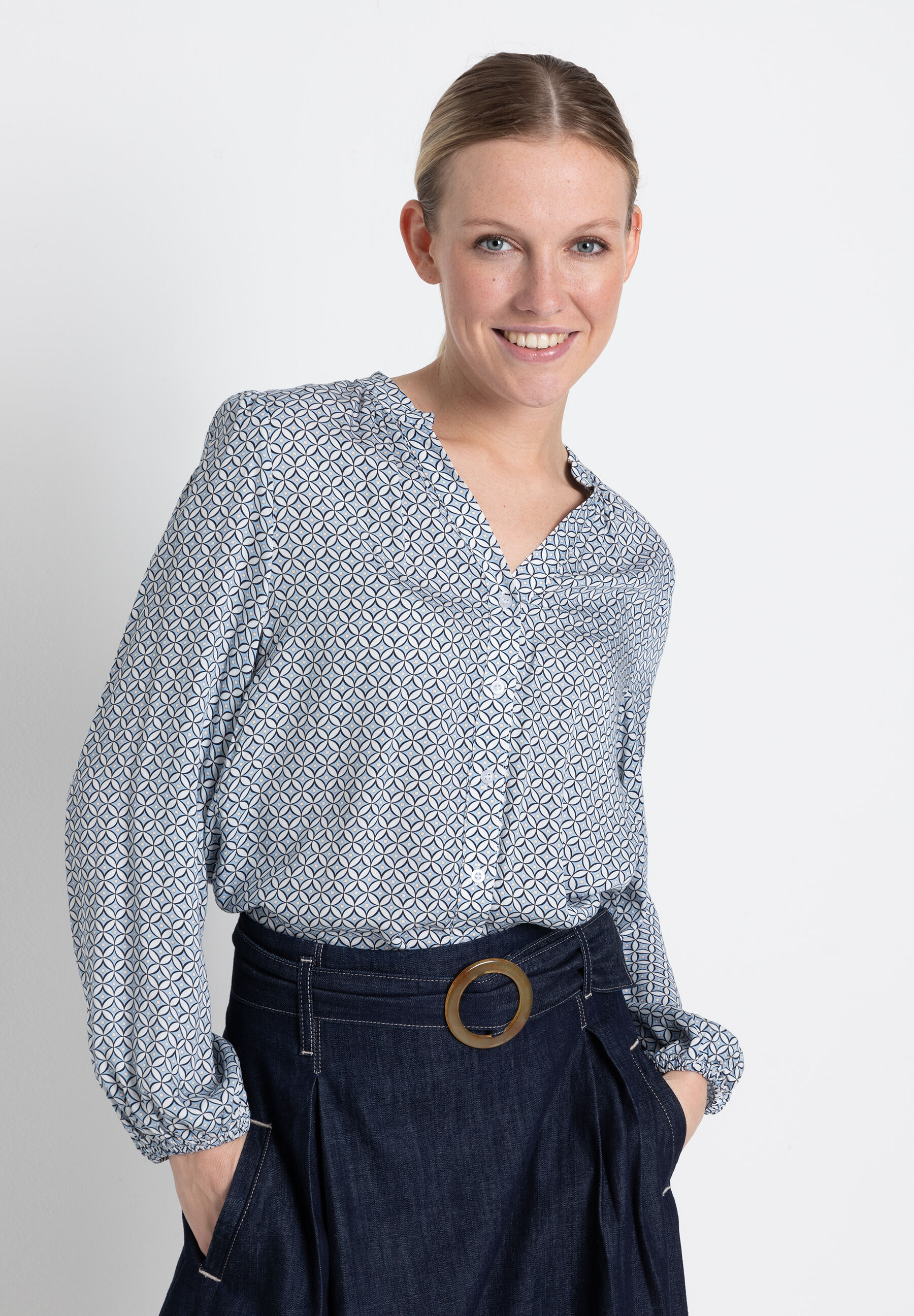 Eine lächelnde Frau in einer fließenden MORE & MORE Viskose-Bluse mit geometrischem Muster und einer dunklen Hose mit Gürtel.