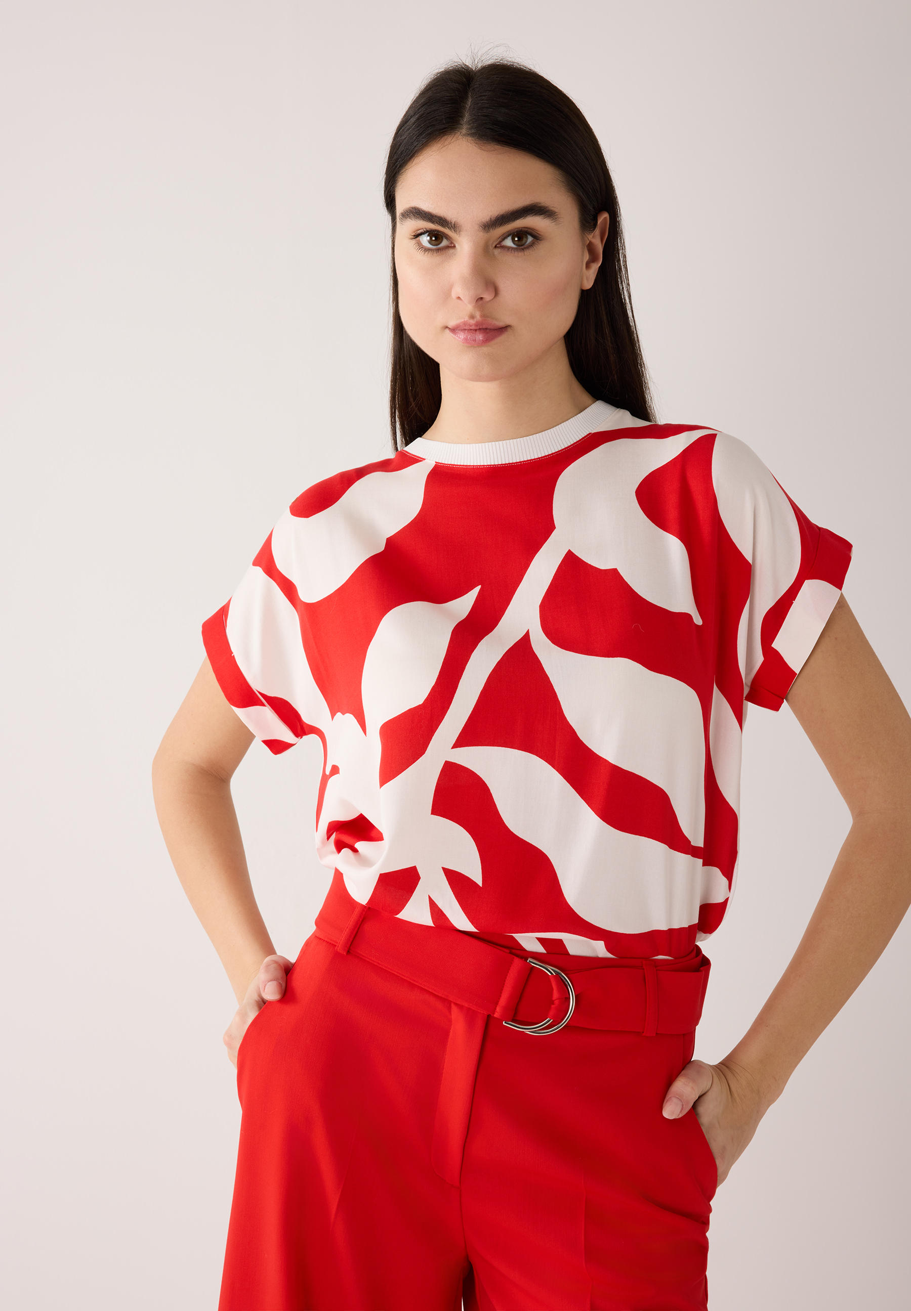 Eine Frau in roter Hose mit passendem Gürtel und einem MORE & MORE Top mit großem, abstrakten Muster.