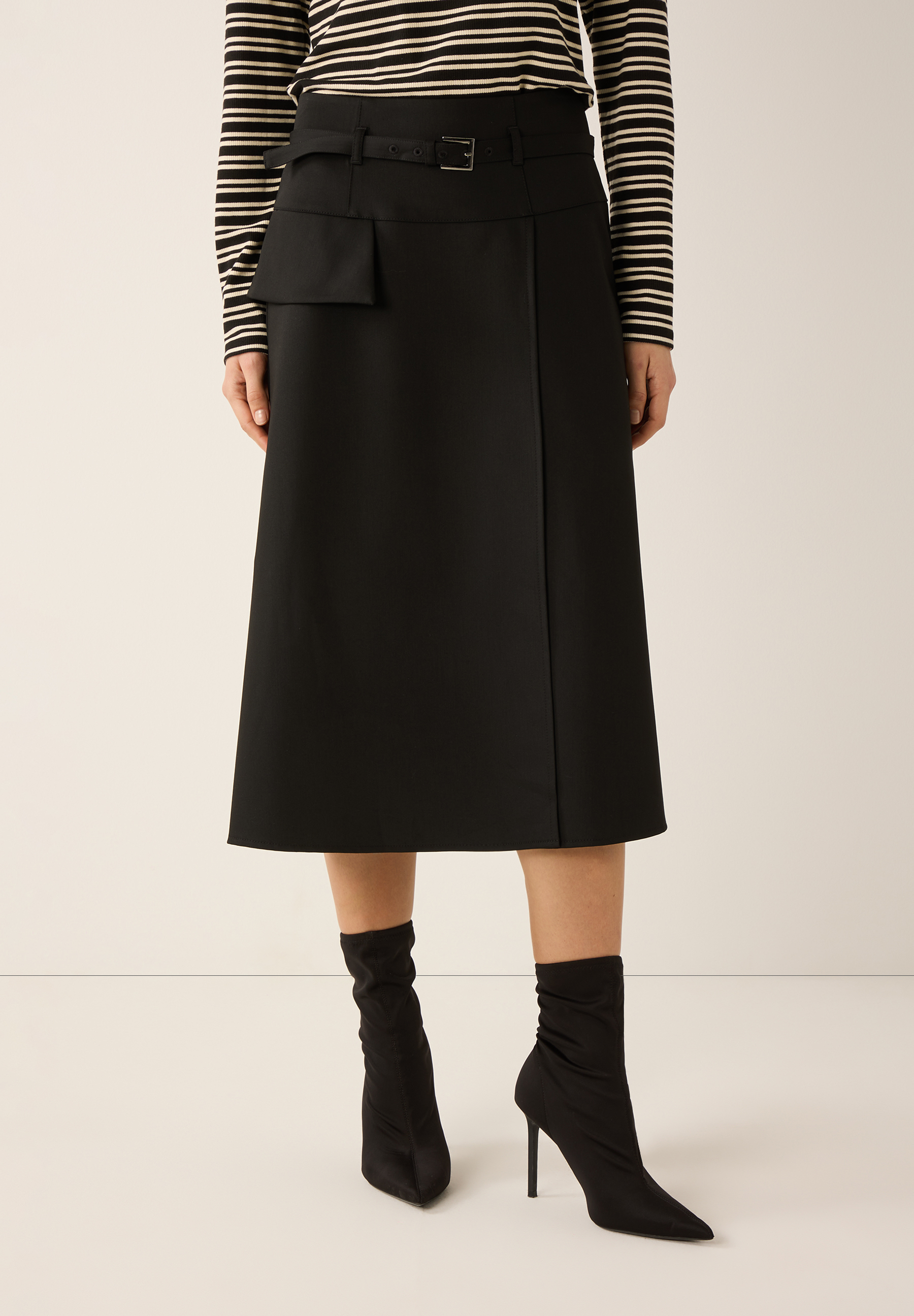 Ein schwarzer MORE & MORE Rock in A-Linie mit Gürtel und Tasche, kombiniert mit einem gestreiften Oberteil und Stiefeln.