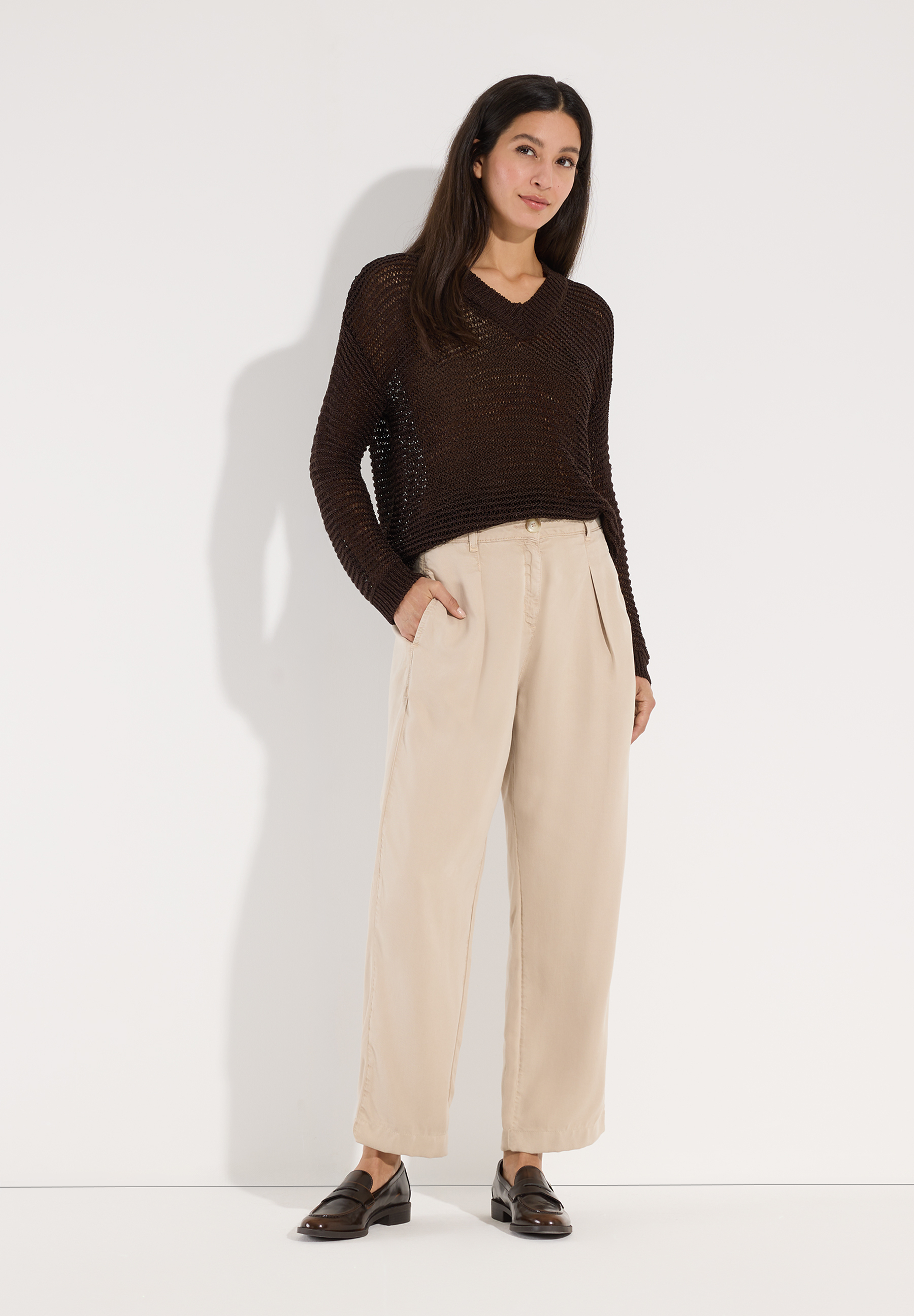 MORE & MORE Pullover: Eine Frau trägt einen braunen Strickpullover mit V-Ausschnitt und beigefarbene Hosen zu Loafers. MORE & MORE Pullover: Eine Frau trägt einen braunen Strickpullover mit V-Ausschnitt und beigefarbene Hosen zu Loafers.