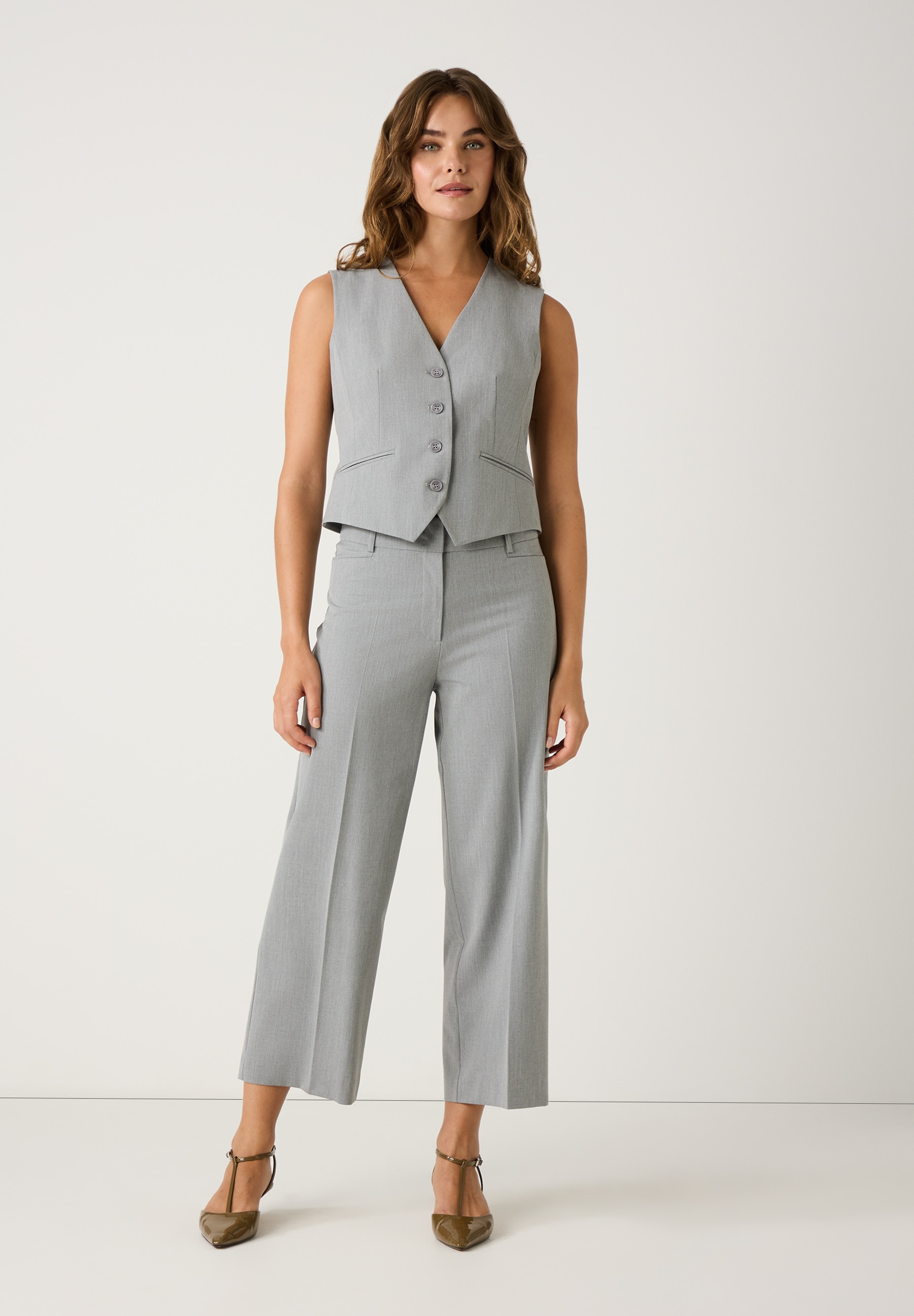 Eine Frau trägt ein graues Hosenanzug-Set von MORE & MORE. Der Blazer ist ärmellos und mit Knöpfen versehen. Das Set besteht aus Viskosemischgewebe. Eine Frau trägt ein graues Hosenanzug-Set von MORE & MORE. Der Blazer ist ärmellos und mit Knöpfen versehen. Das Set besteht aus Viskosemischgewebe.