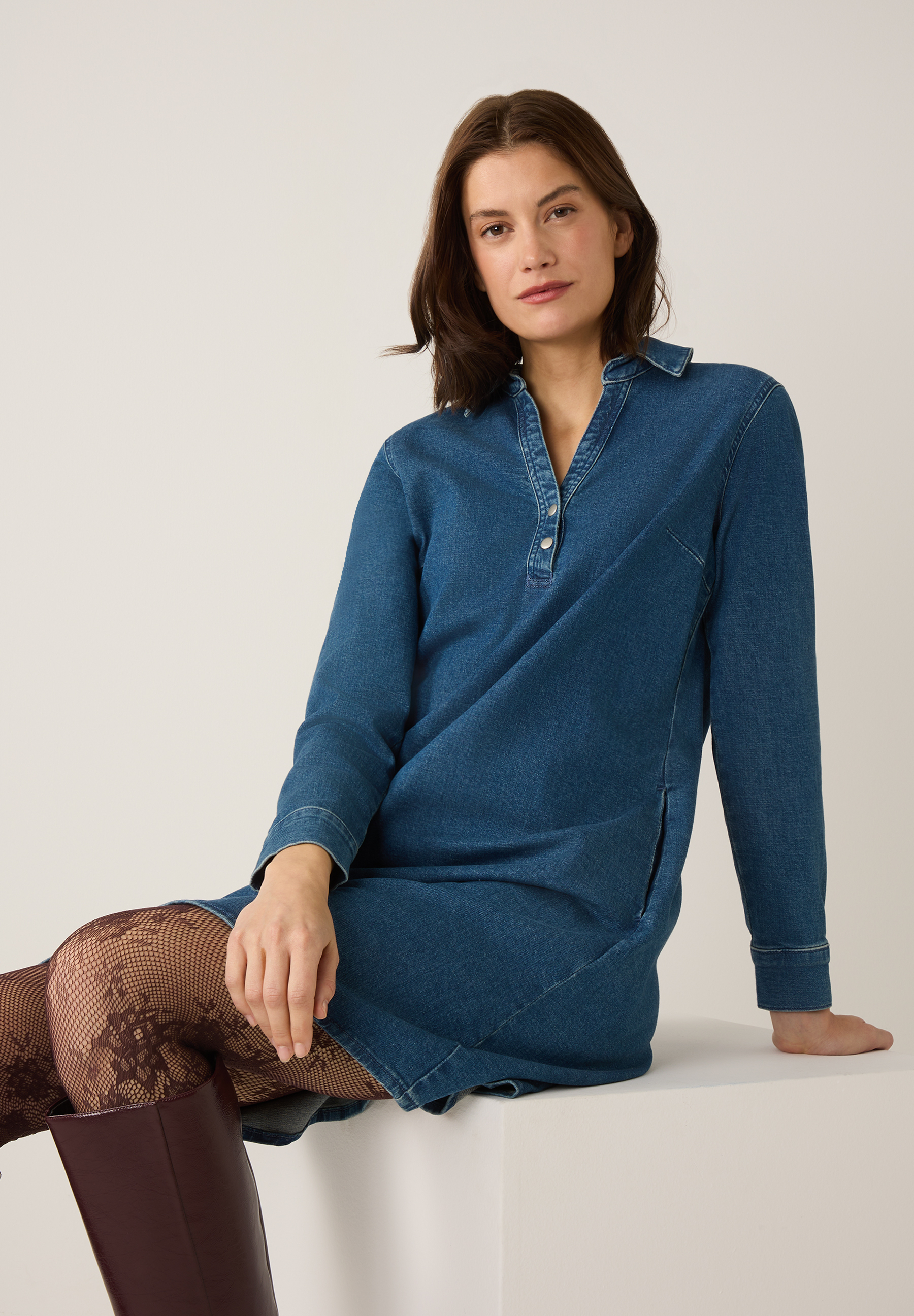 Eine Frau trägt ein Jeanskleid der Marke MORE & MORE, dazu blickdichte Strumpfhose und Stiefel.