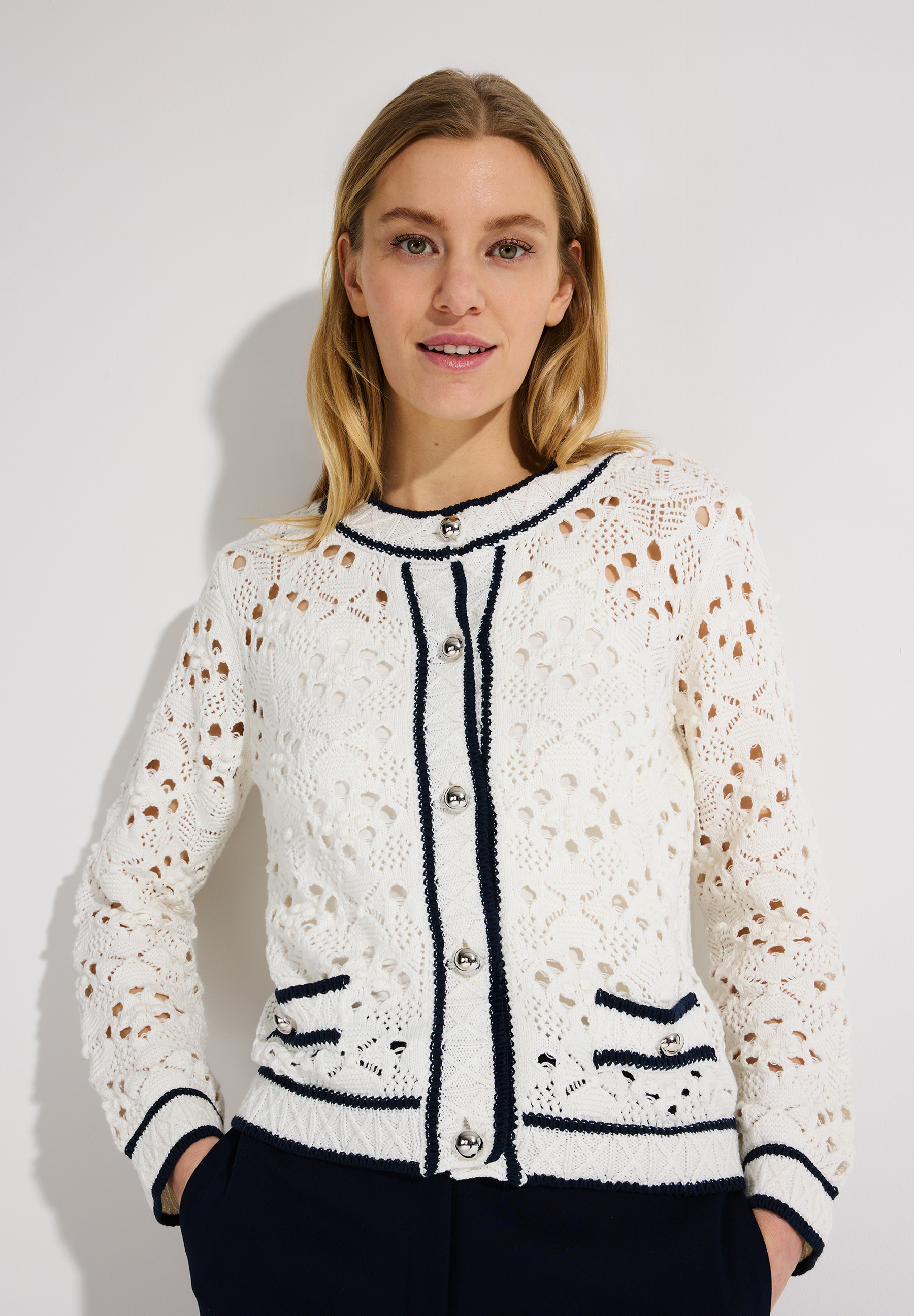 Eine Frau mit einem MORE & MORE Lochstrick-Oberteil. Die Strickjacke ist weiß mit Knöpfen und blauen Akzenten.