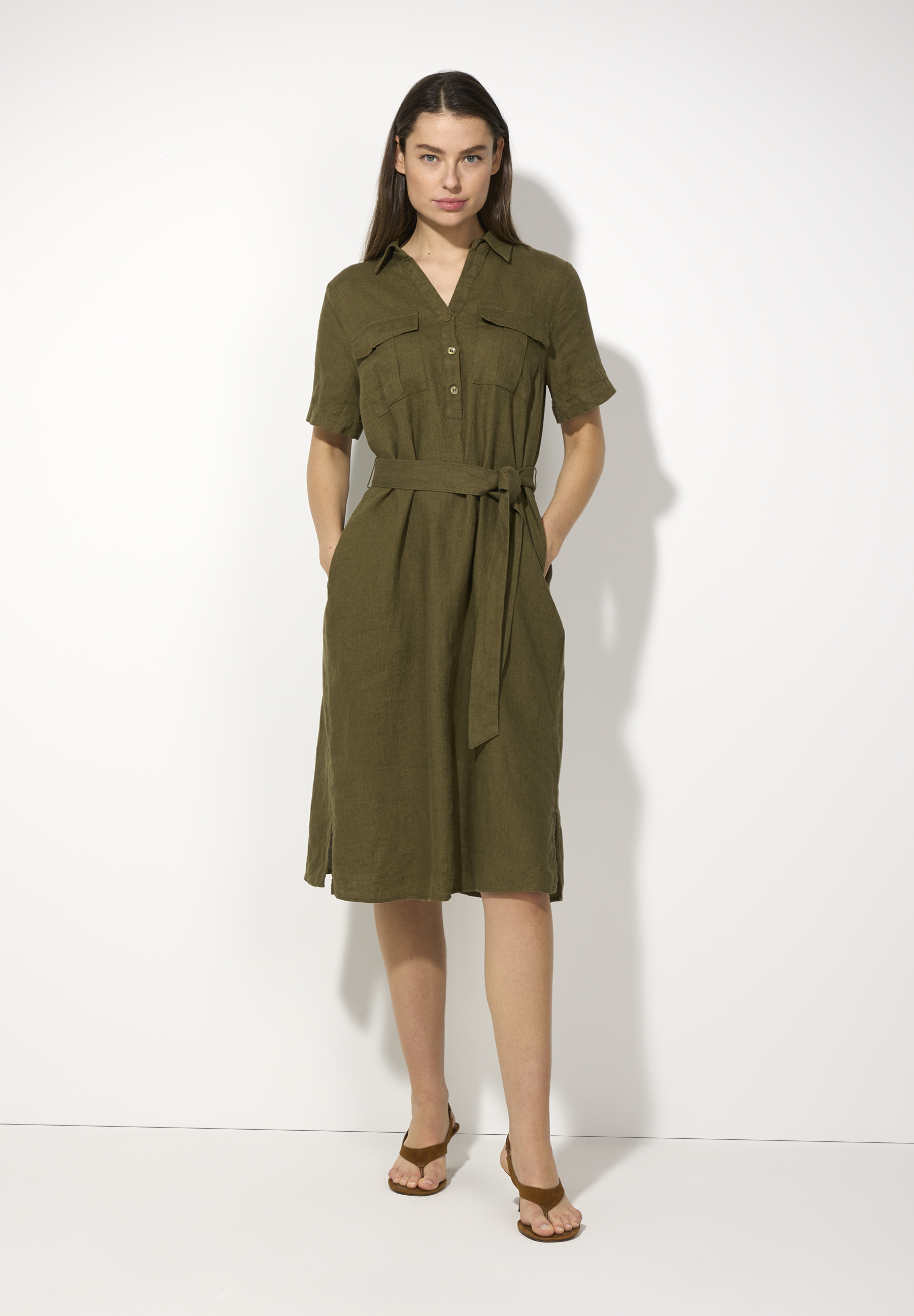 Eine Frau trägt ein olivgrünes MORE & MORE Leinenkleid mit Gürtel und Sandalen vor einem weißen Hintergrund.