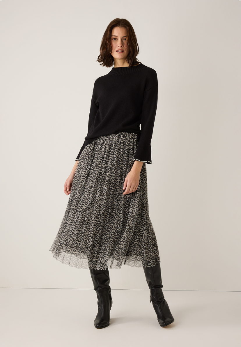 Eine Frau trägt einen schwarzen MORE & MORE Strickpullover, einen gemusterten Rock und Stiefel. Eine Frau trägt einen schwarzen MORE & MORE Strickpullover, einen gemusterten Rock und Stiefel.