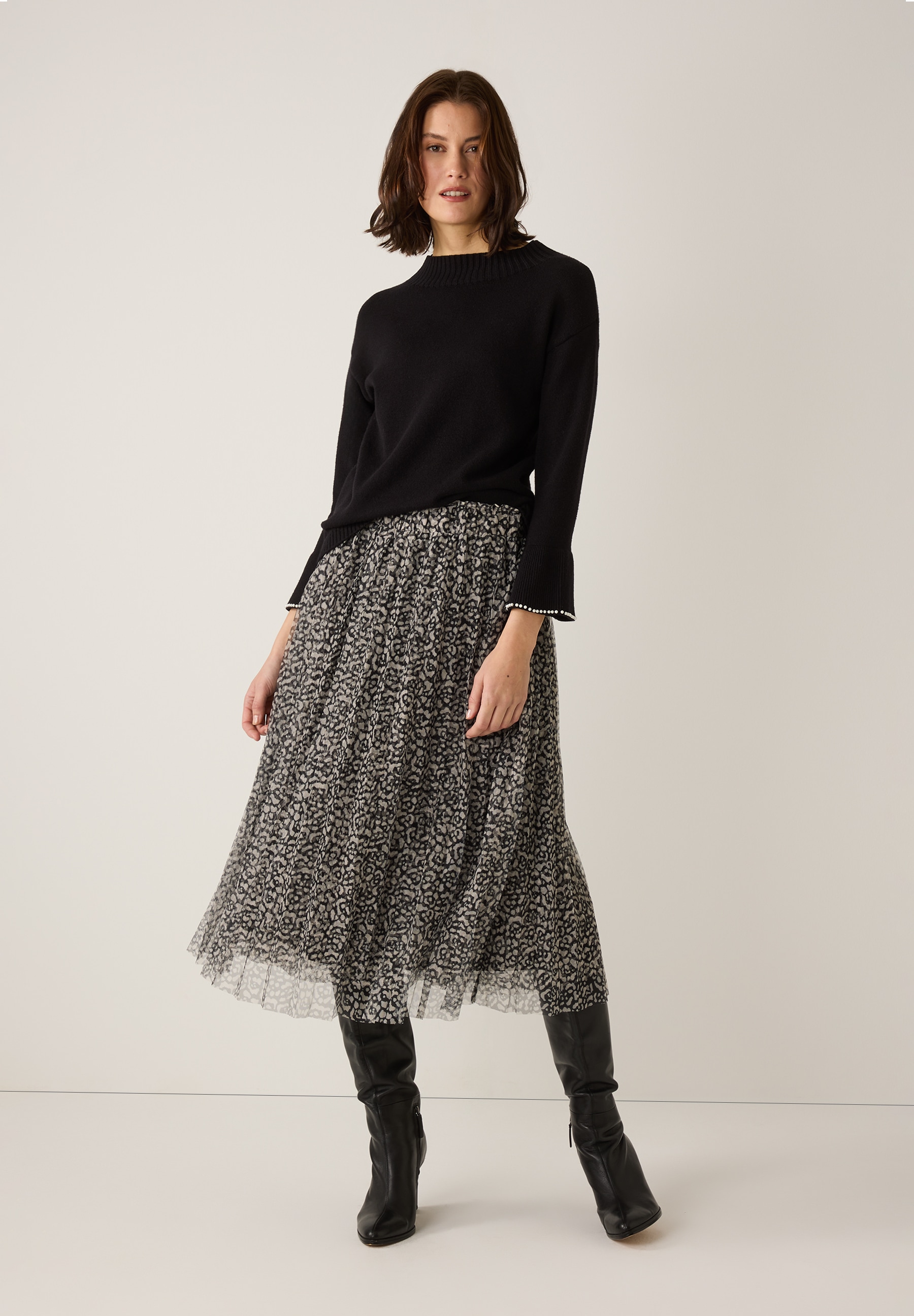 Eine Frau trägt einen schwarzen MORE & MORE Strickpullover, einen gemusterten Rock und Stiefel. Eine Frau trägt einen schwarzen MORE & MORE Strickpullover, einen gemusterten Rock und Stiefel.