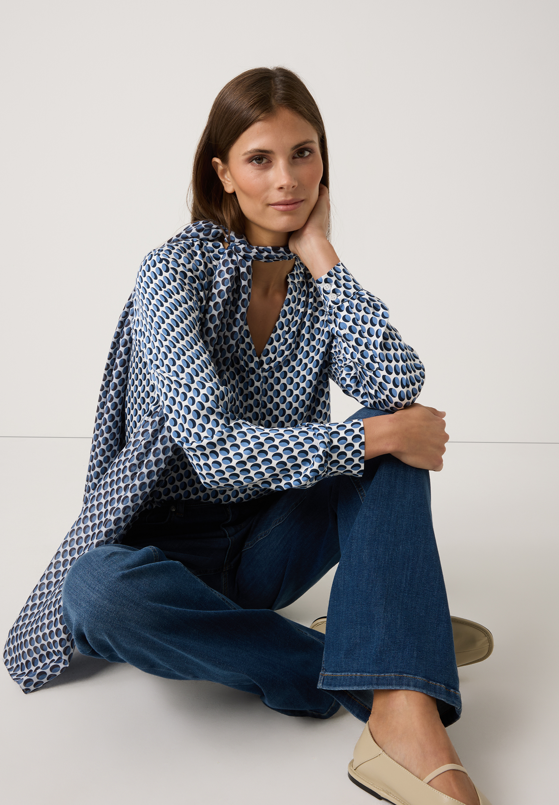 Eine Frau sitzt auf dem Boden und trägt eine blaue MORE & MORE Bluse mit Punktmuster und Jeans. Eine Frau sitzt auf dem Boden und trägt eine blaue MORE & MORE Bluse mit Punktmuster und Jeans.