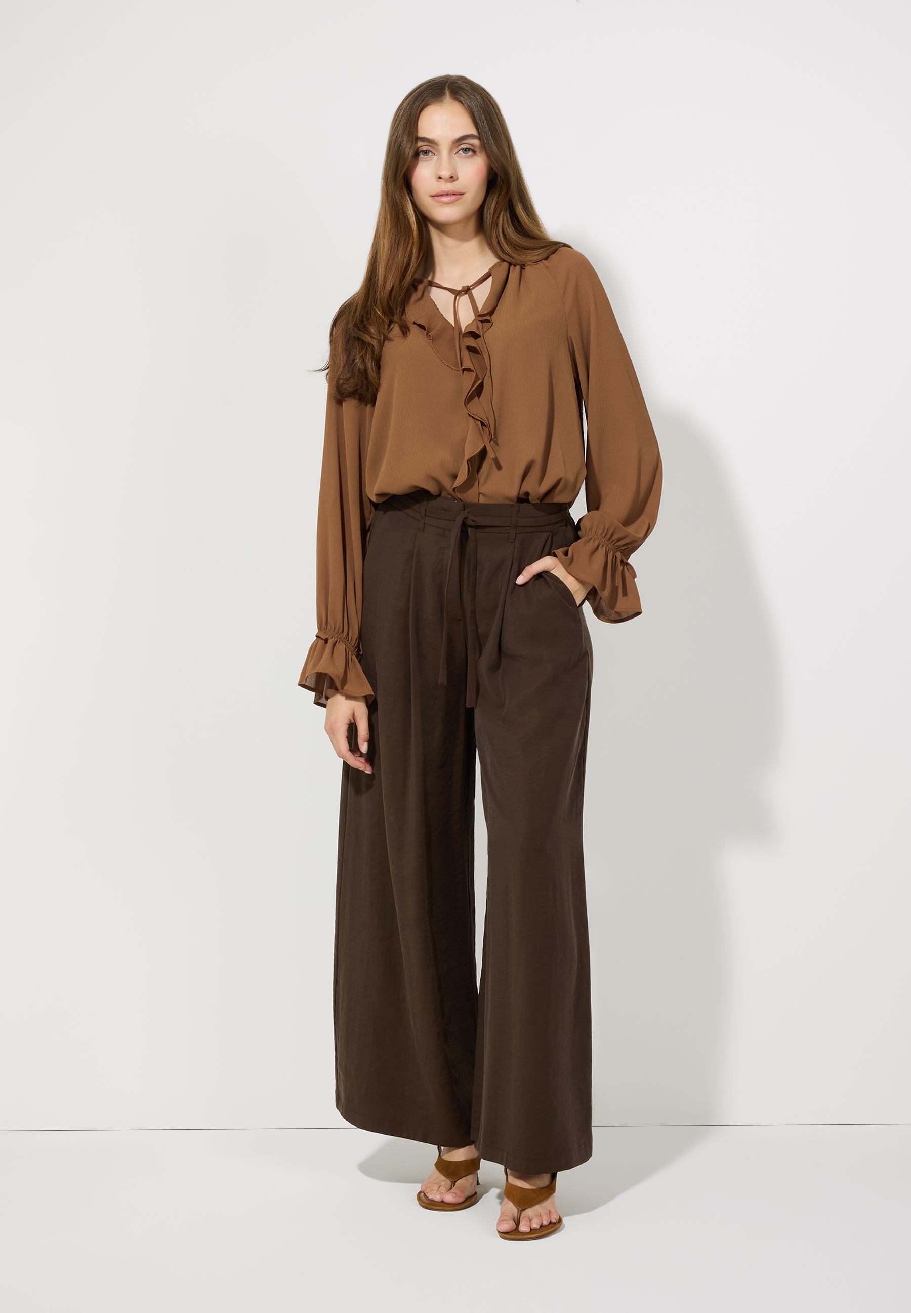Eine Frau im braunen MORE & MORE-Ensemble: Bluse mit Rüschen und weite Hose. Sie posiert vor einem weißen Hintergrund. Eine Frau im braunen MORE & MORE-Ensemble: Bluse mit Rüschen und weite Hose. Sie posiert vor einem weißen Hintergrund.