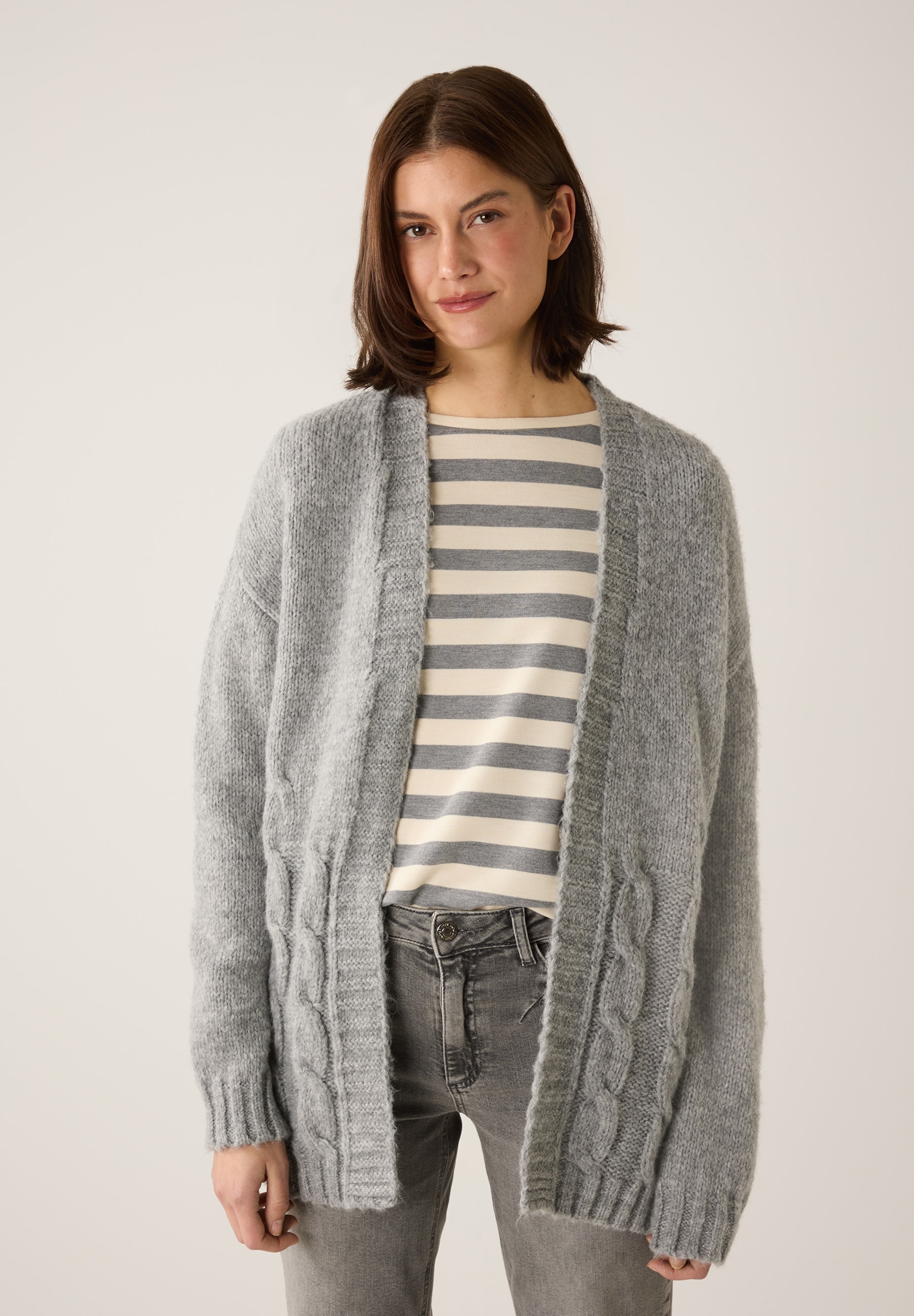 Eine Frau trägt eine Strickjacke von MORE & MORE über einem gestreiften Oberteil und graue Jeans. Eine Frau trägt eine Strickjacke von MORE & MORE über einem gestreiften Oberteil und graue Jeans.