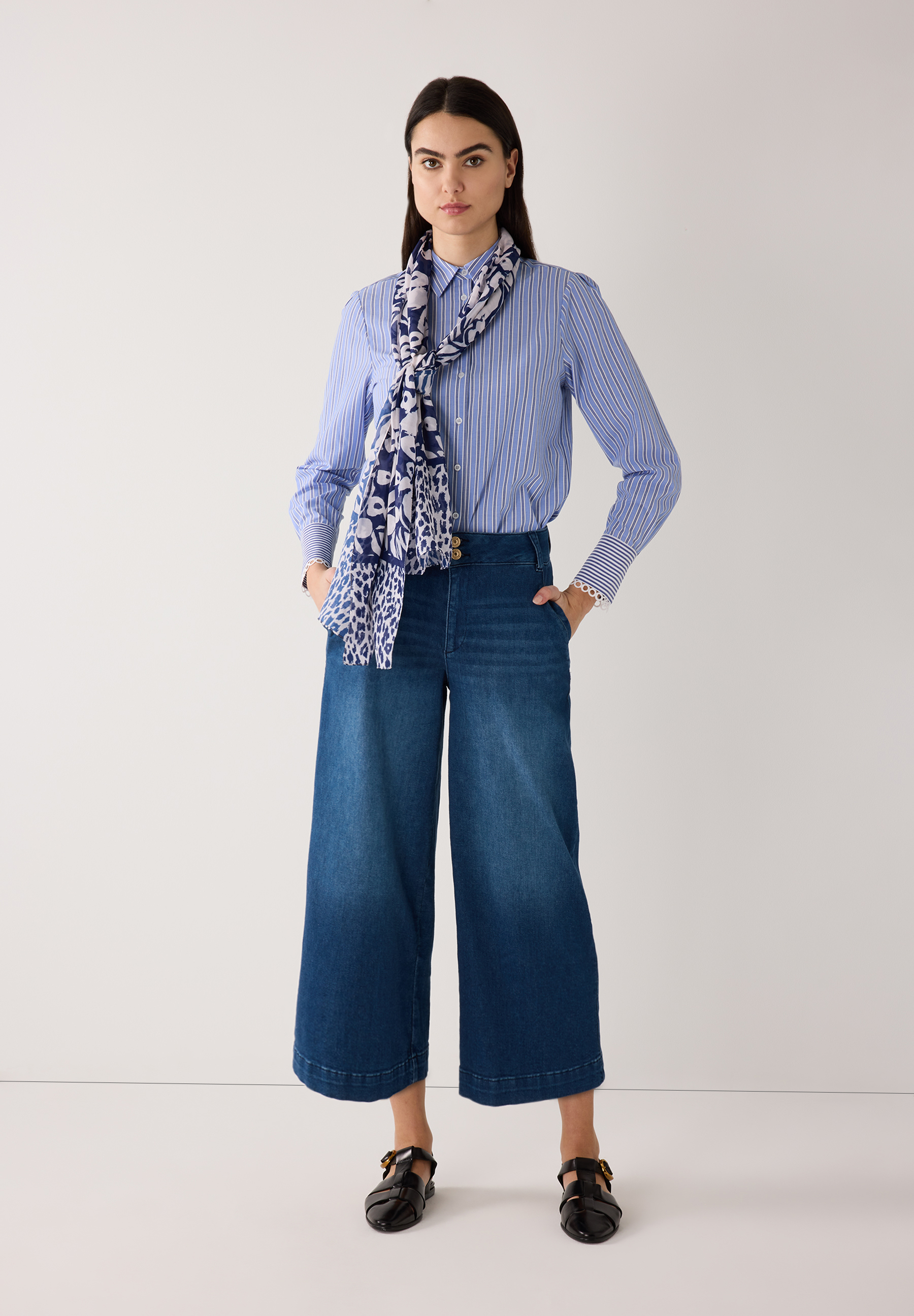 Eine Frau in MORE & MORE-Popeline-Bluse mit Schal und Jeans-Culottes posiert vor einem weißen Hintergrund. Eine Frau in MORE & MORE-Popeline-Bluse mit Schal und Jeans-Culottes posiert vor einem weißen Hintergrund.