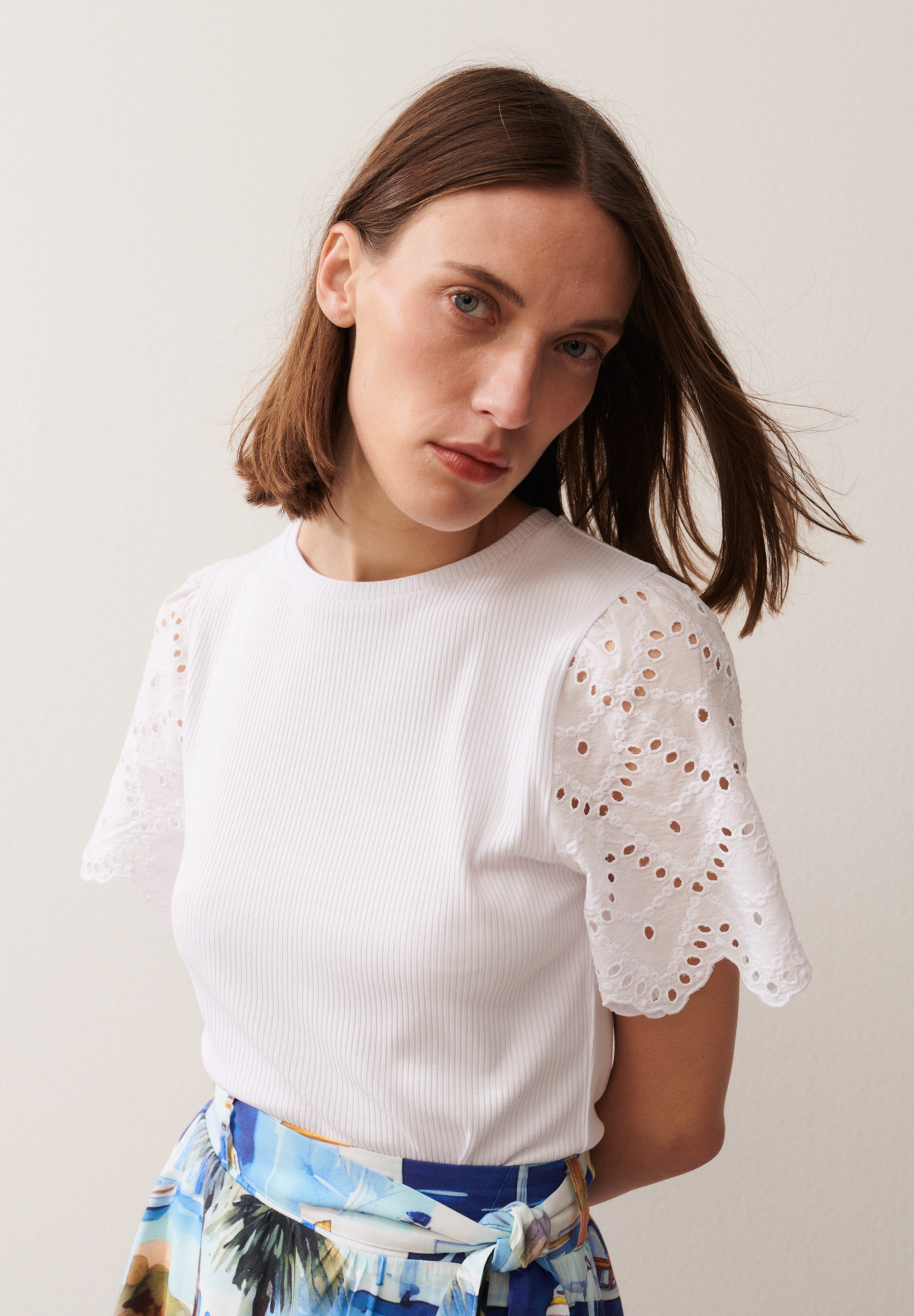 Eine Frau trägt ein geripptes, weißes MORE & MORE Oberteil mit Lochstickerei-Ärmeln und einen Rock mit blauem Muster. Eine Frau trägt ein geripptes, weißes MORE & MORE Oberteil mit Lochstickerei-Ärmeln und einen Rock mit blauem Muster.