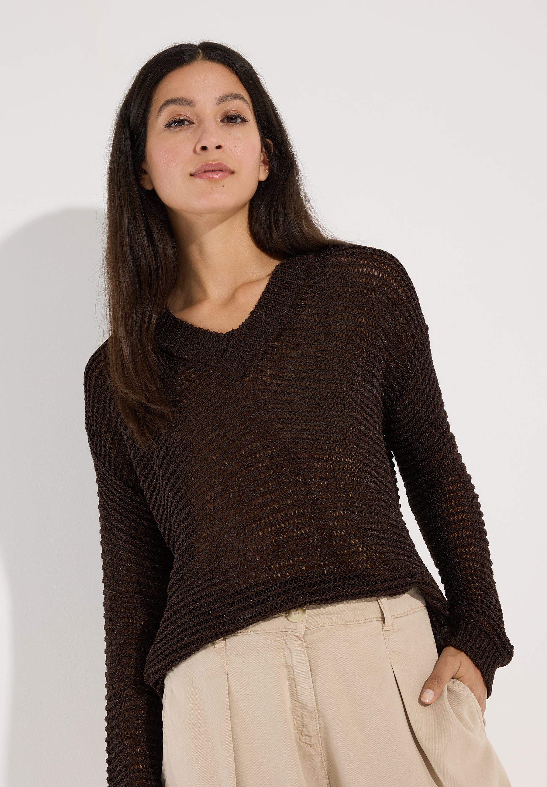 MORE & MORE Damen Pullover: Nahaufnahme einer Frau in einem braunen V-Ausschnitt Strickpullover und beiger Hose, Hand in der Hosentasche.