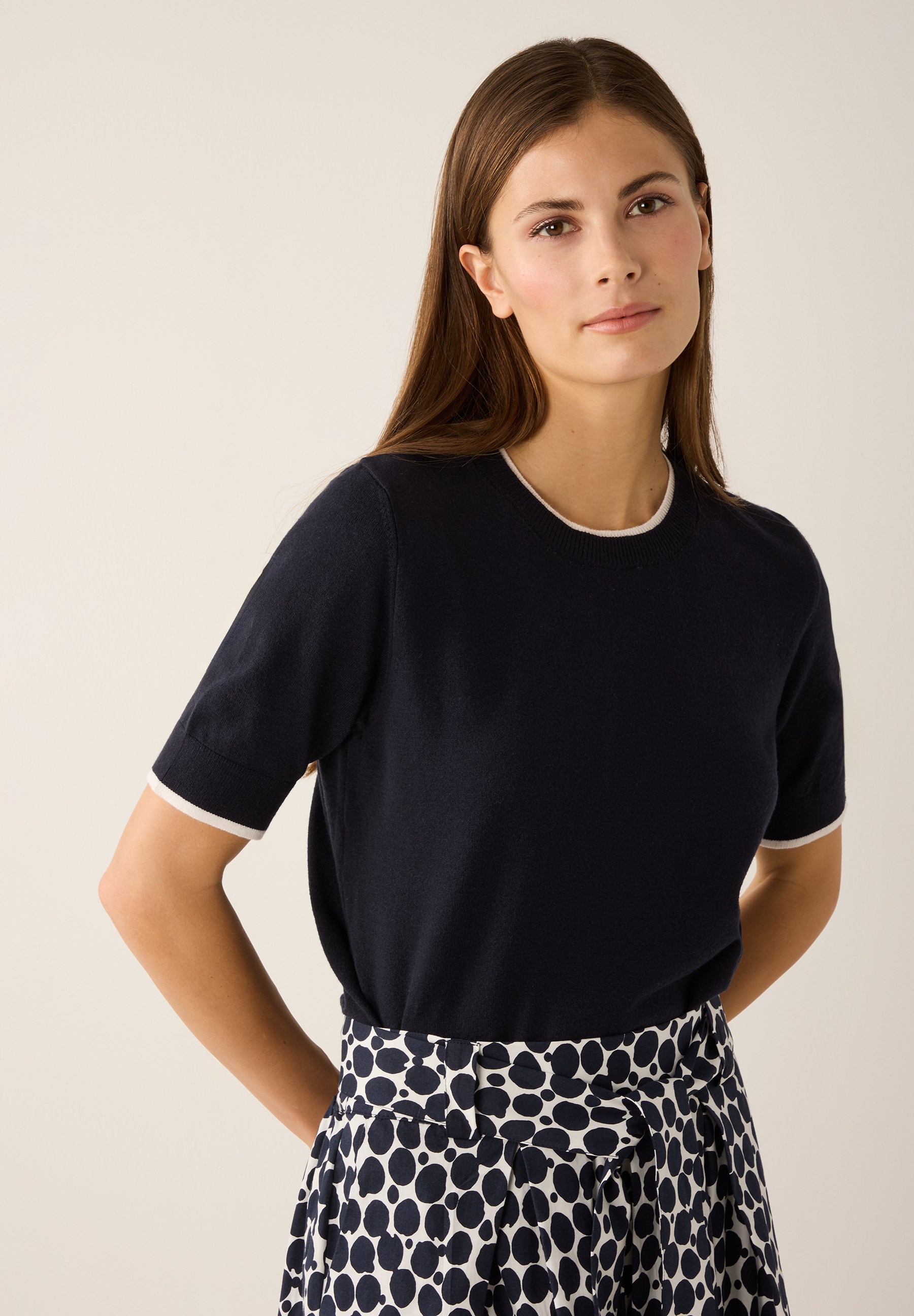 Eine Frau trägt ein marineblaues Feinstrick-Oberteil von MORE & MORE mit weißen Akzenten an Kragen und Ärmeln. Eine Frau trägt ein marineblaues Feinstrick-Oberteil von MORE & MORE mit weißen Akzenten an Kragen und Ärmeln.