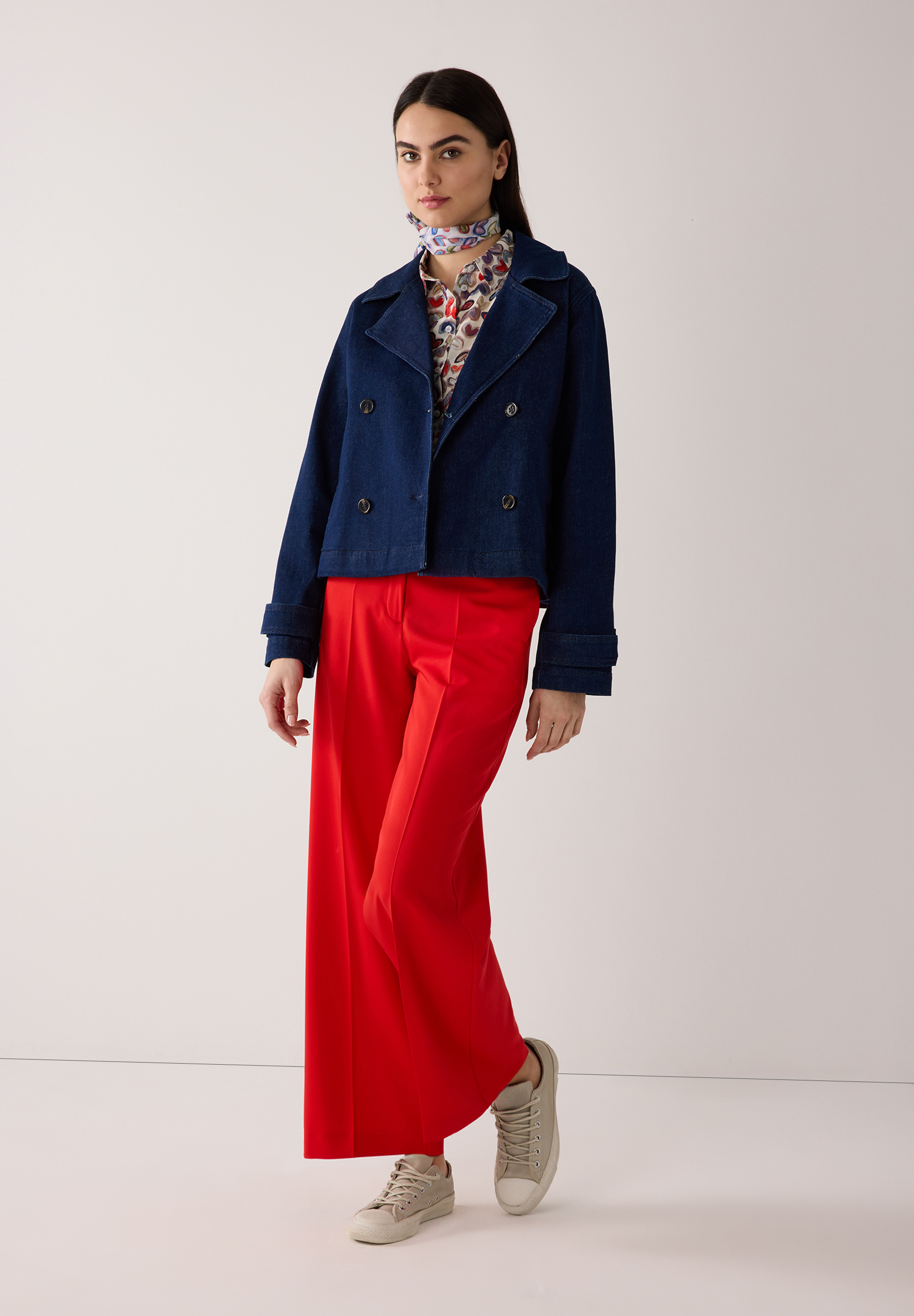Eine Frau in MORE & MORE Oberteilen: Jeansjacke, gemustertes Hemd, roter Hose und beigen Sneakers vor einem weißen Hintergrund.