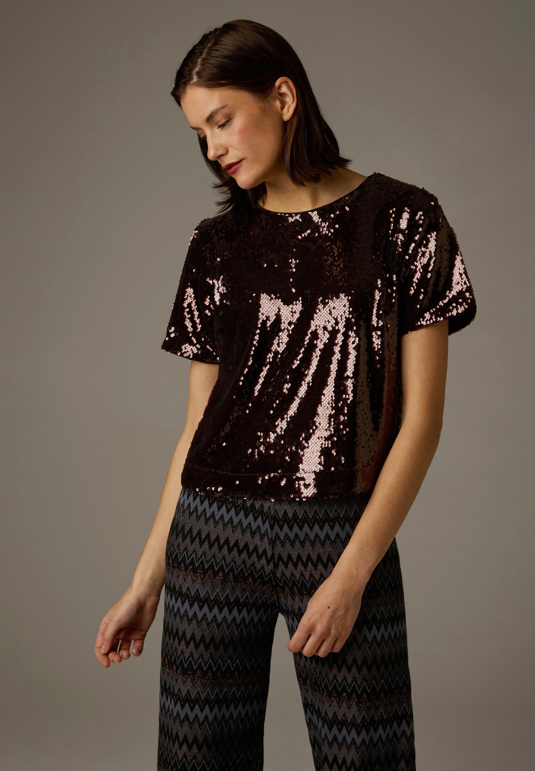 Eine Frau mit verschränkten Armen trägt ein glänzendes MORE & MORE Oberteil aus Polyester mit Pailletten.